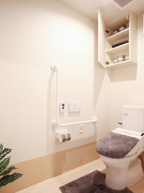 白で統一された明るい空間で便座カバーとトイレマットが設置されている。また、予備のトイレットペーパーは棚に保管されている。
