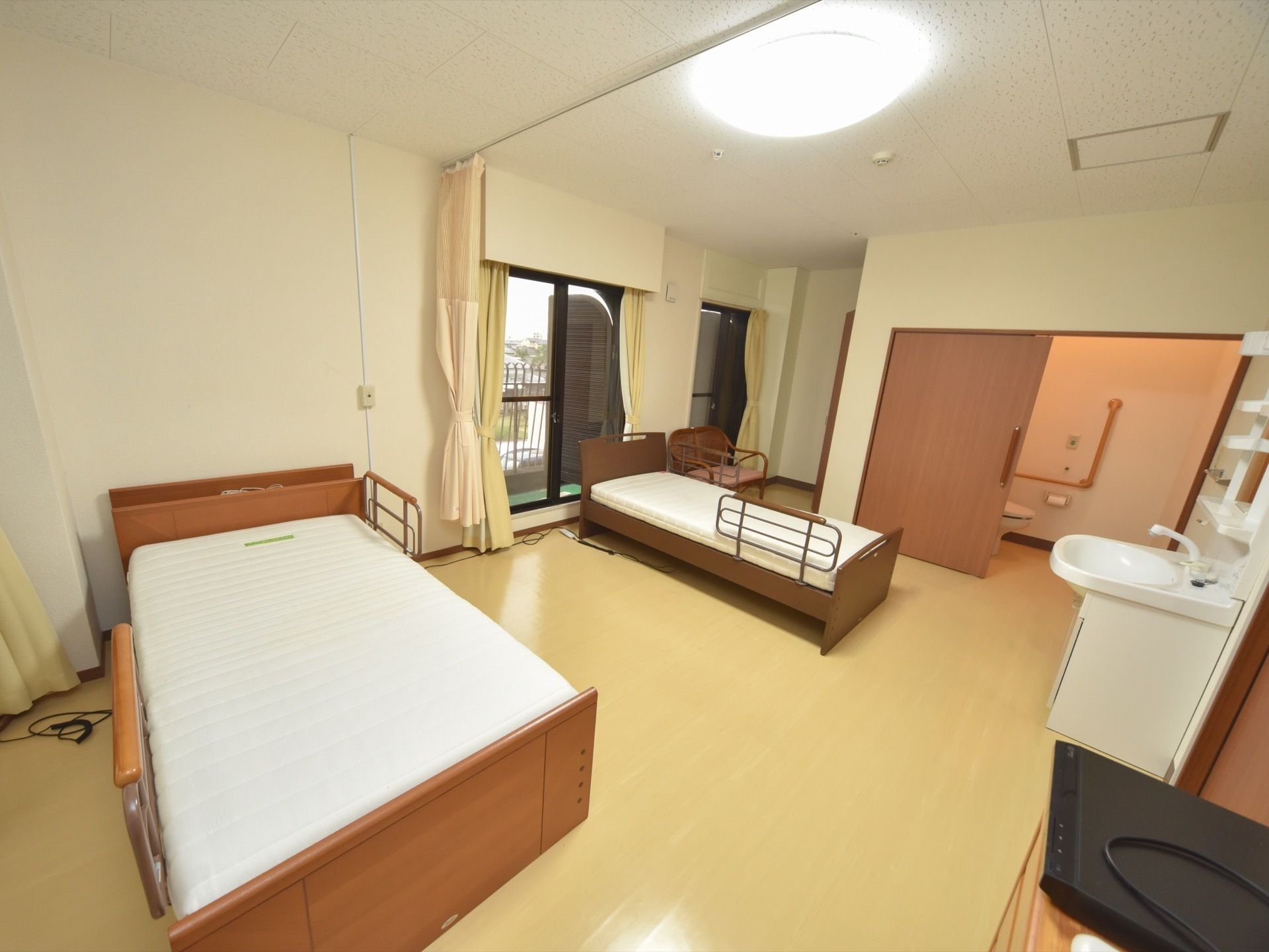広々とした二人部屋の居室には、介護用ベッドが置かれている。また室内には窓があり、カーテンが取り付けられている。