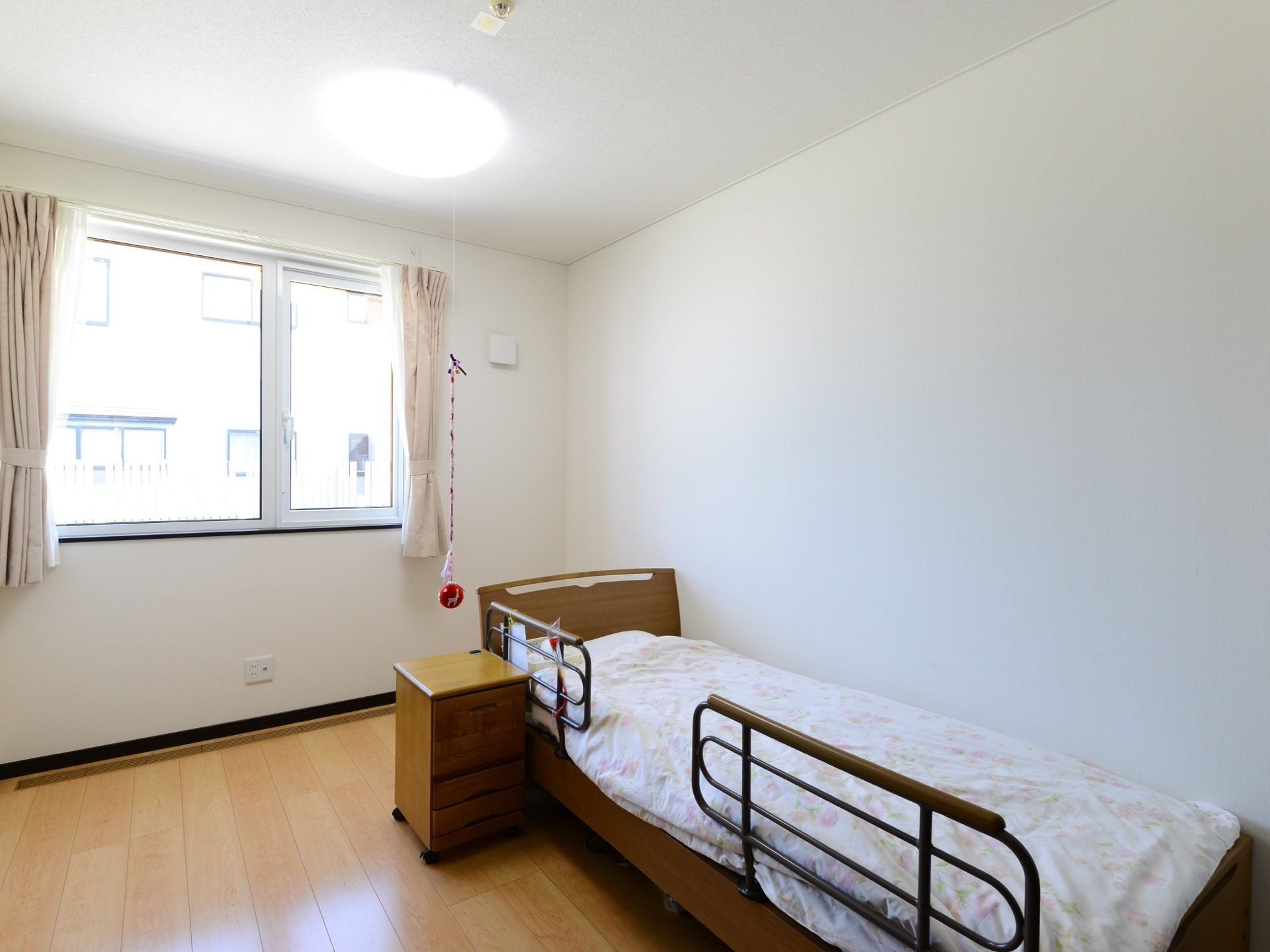 日当たりが良いワンルームの居室である。窓にはカーテンが掛けられており、ベッドには寝具が用意されている。