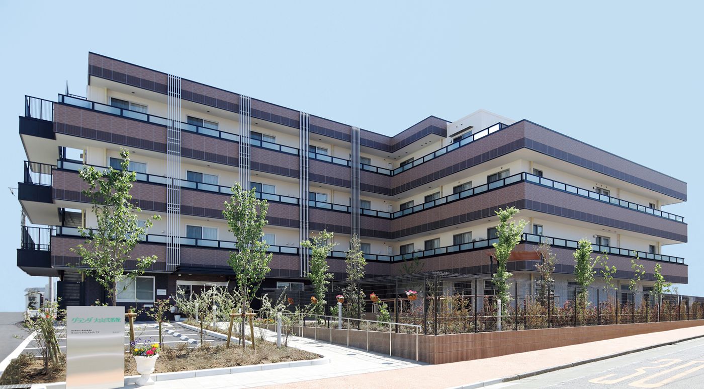 手すりがついた長いスロープのある玄関と、ピンク色の外観が温かみを感じさせる４階建てのマンションのような建物