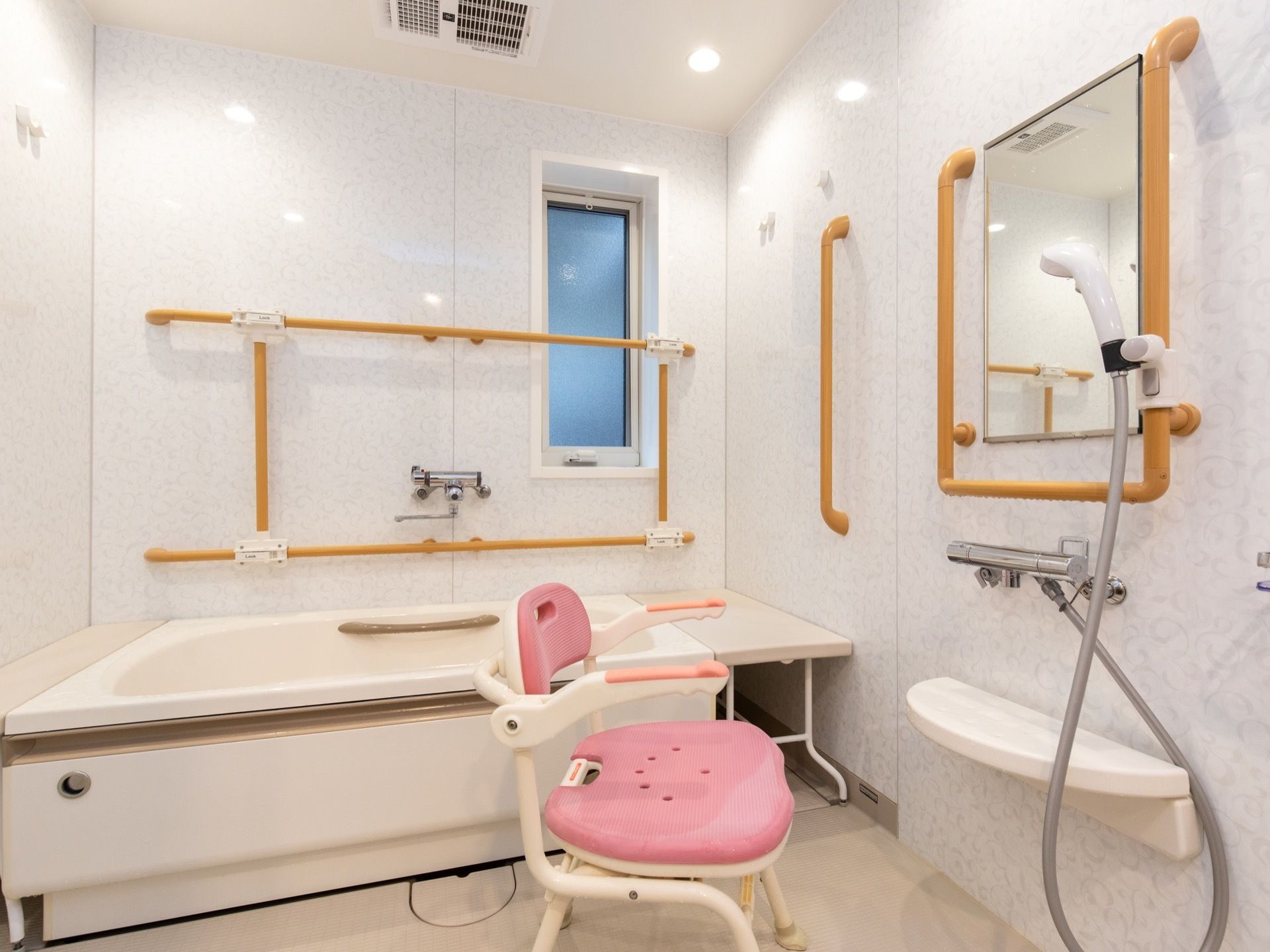 浴室内は白を基調とした内装である。濃いベージュ色の手すりと、シャワーチェアのピンクの座面がアクセントになっている。