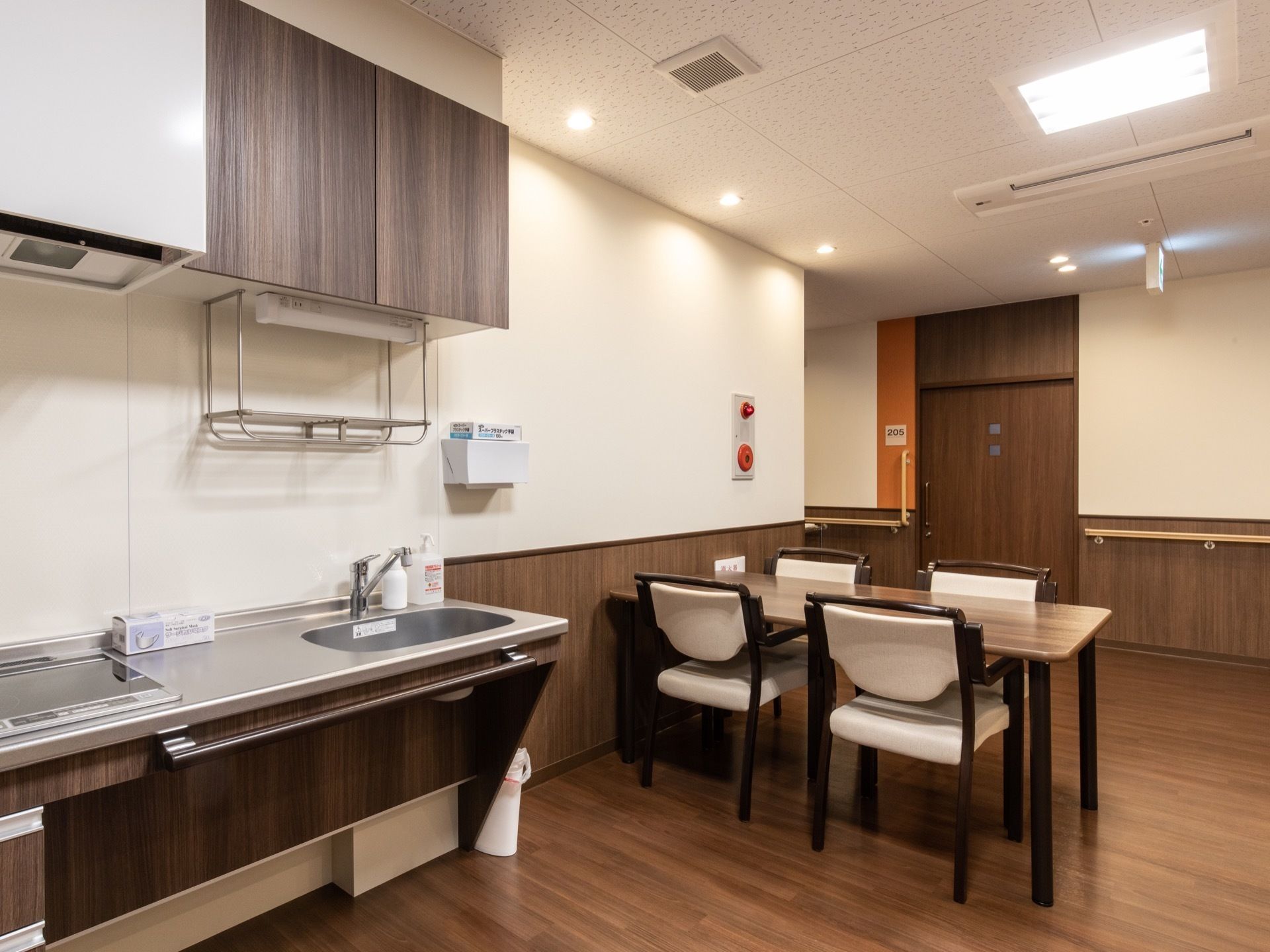 共有スペースにシステムキッチンとダイニングセットがある。キッチンはダークブラウンの木目調で、シンクの手前にタオル掛けにもなる手すりがある。