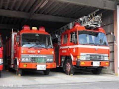 広い倉庫のような駐車スペースの中に、はしご車など種類が違う消防車が複数台駐車されている。地面はアスファルトで舗装されている。