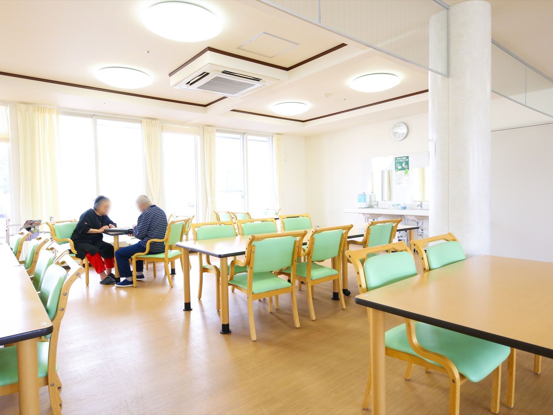 食堂は、大きな窓からの採光や天井の照明で明るくなっている。入居者様が２名、テーブルをはさんで座っている。