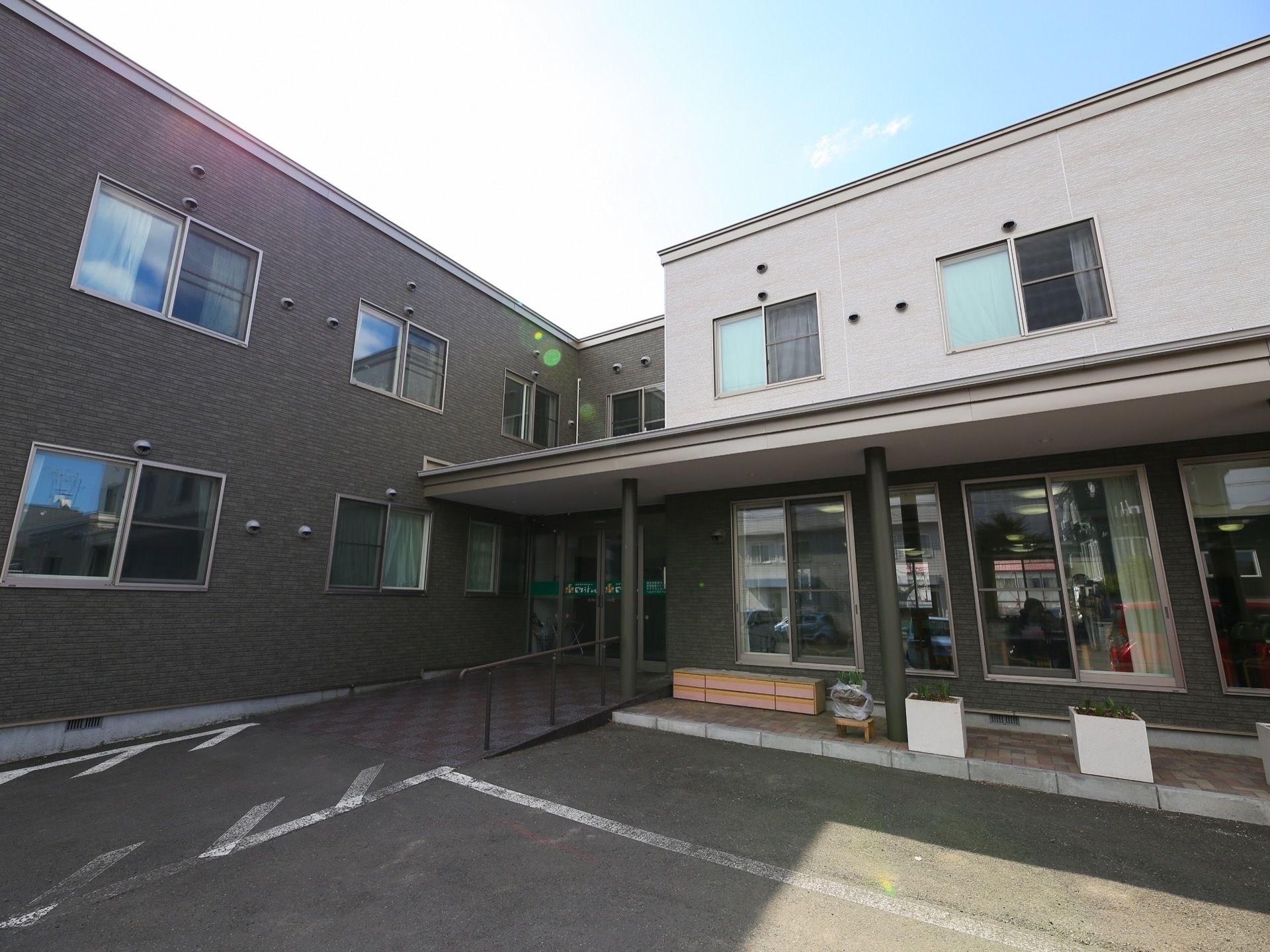 灰色と白色のコントラストの外壁の２階建ての施設である。玄関前には手すりが設置されたゆるやかなスロープがある。
