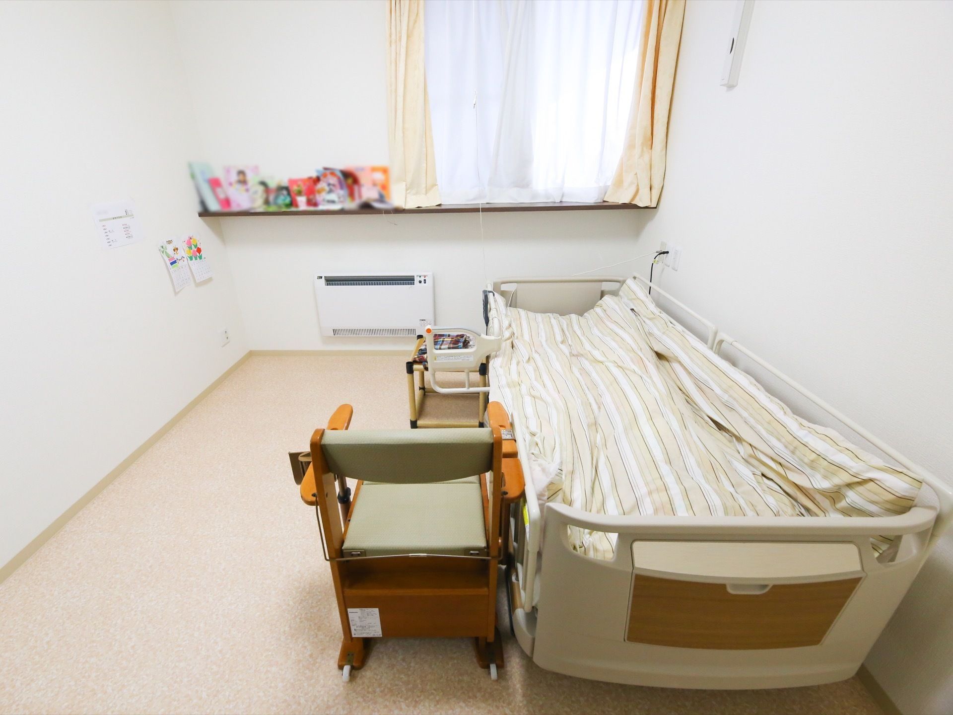 居室の窓際に飾り棚があり、写真や作品が飾られている。ベッドの横には、背もたれやひじ掛けがついた椅子が置かれている。