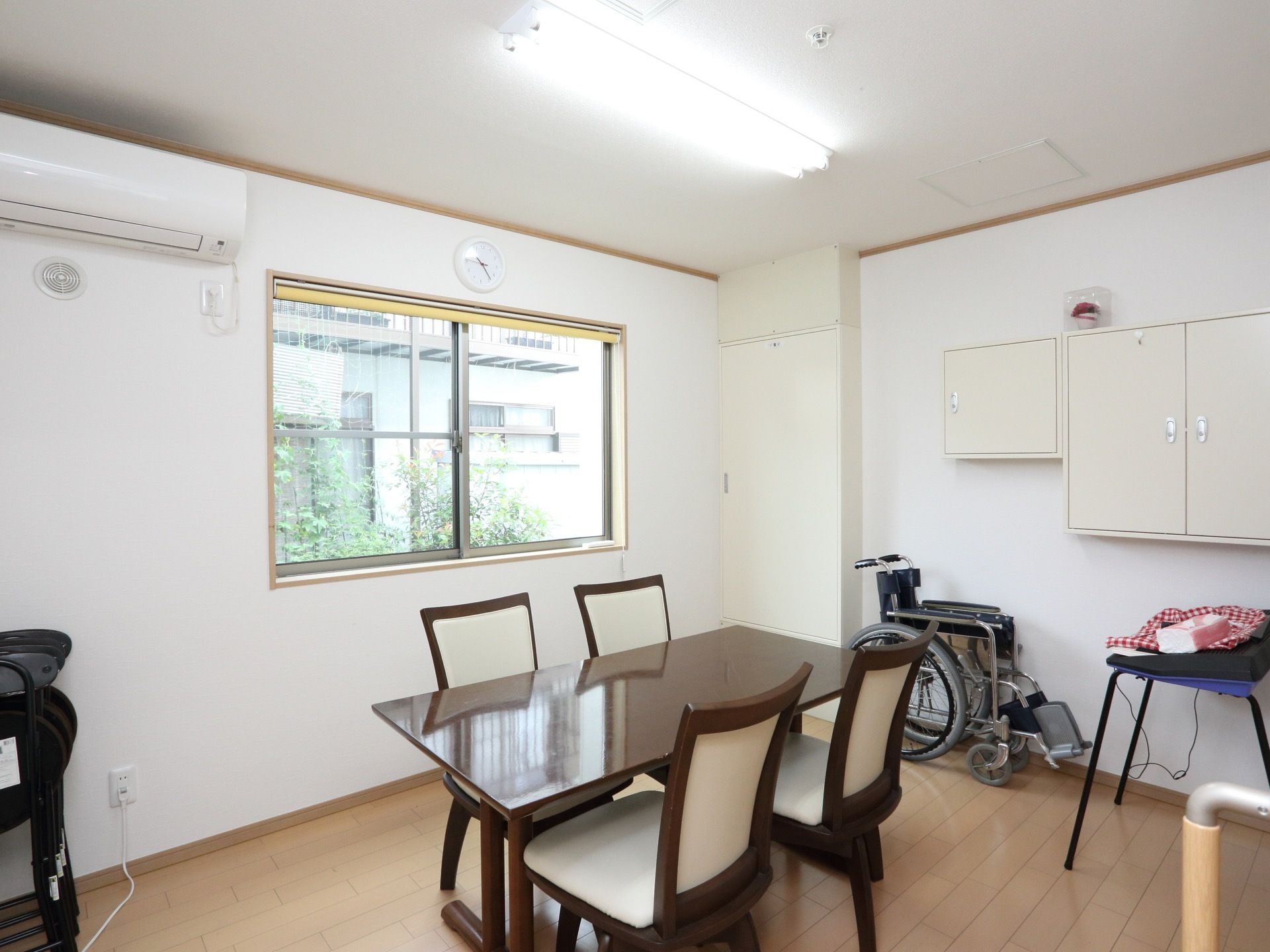 フローリングの床の部屋にテーブルと椅子が４脚設置されている。奥に折りたたまれた車椅子が置かれている。壁にエアコンが取りつけてある。