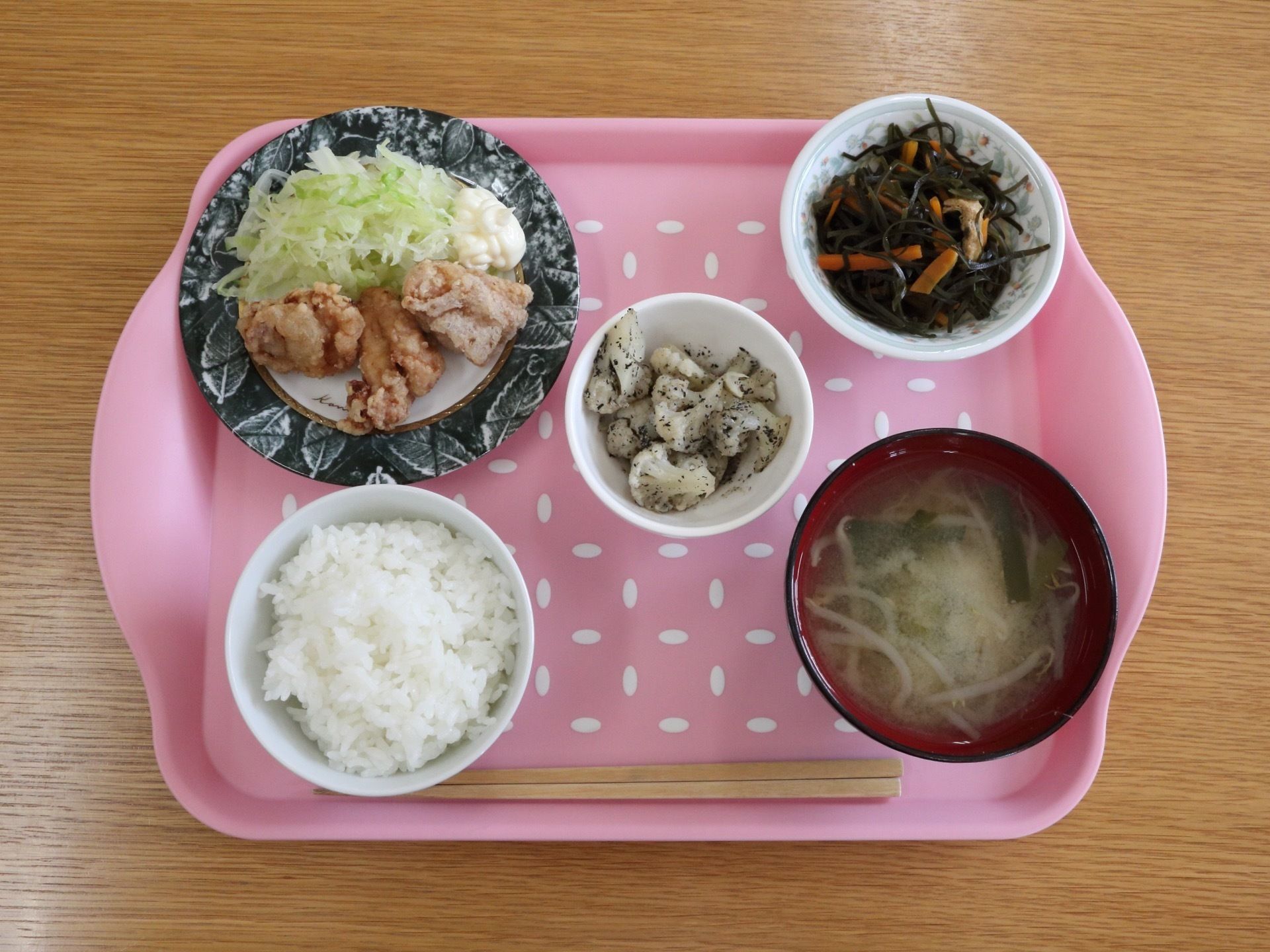 ピンクのトレイにから揚げと野菜の添えられた皿とひじきなどの入った小皿が２皿に、ごはんと味噌汁のお椀が並んでいる。