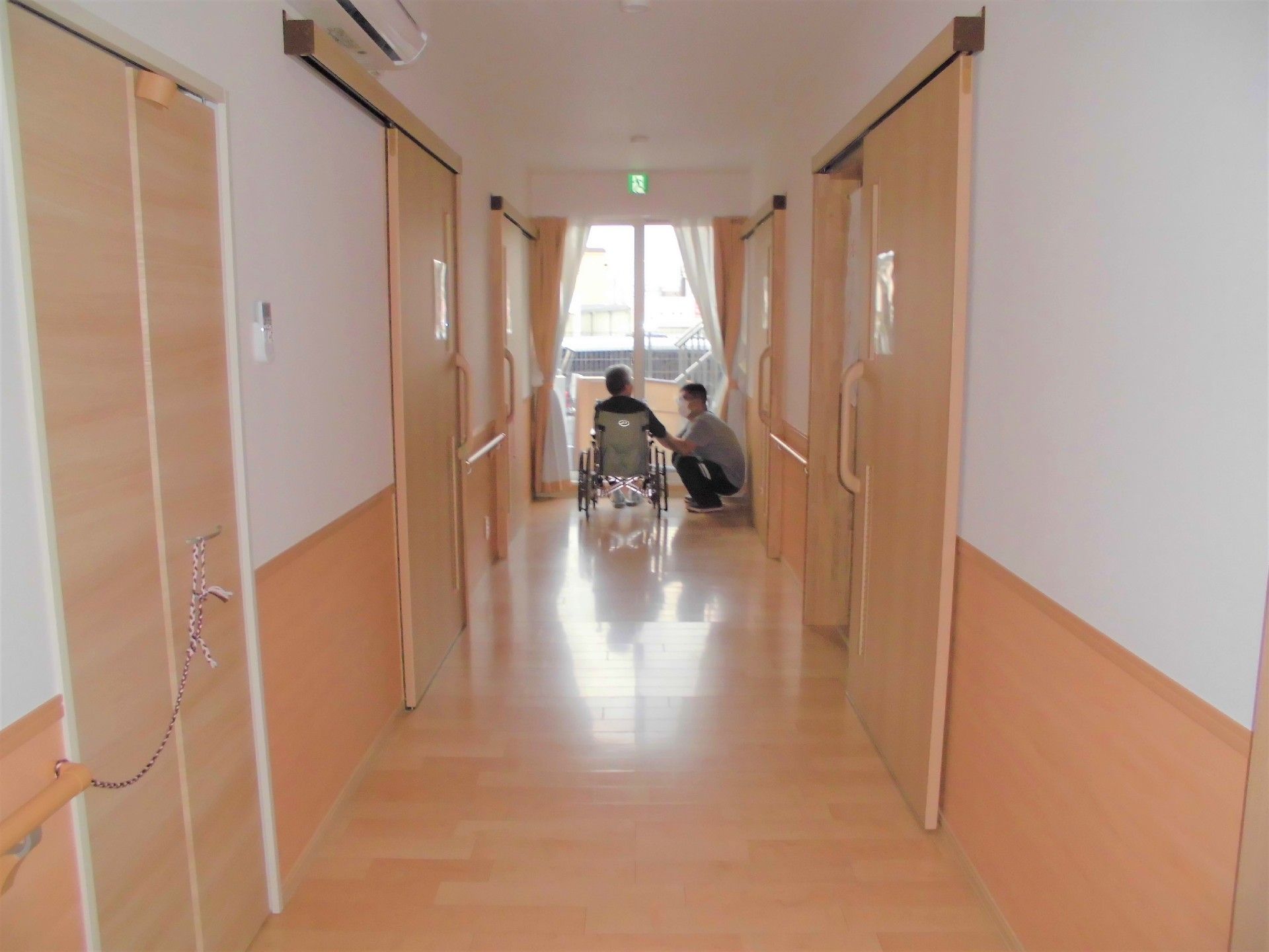 まっすぐに続く廊下とその向こうにいる高齢者を写したもの。両側に個室の入り口がある様子