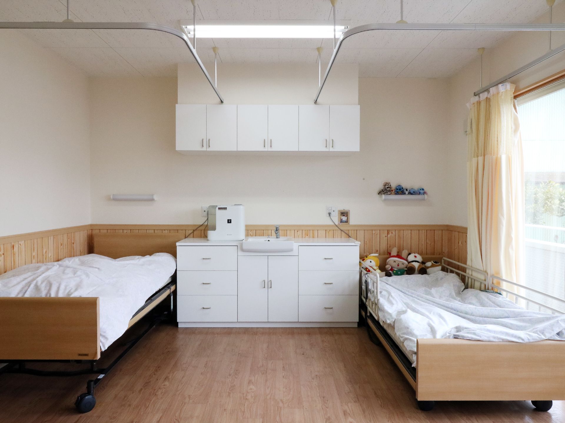 白とベージュの壁が明るい雰囲気の二人部屋。収納に便利な吊戸棚と洗面台付チェストが備え付けてある。ベッドはカーテンで間仕切り可能。