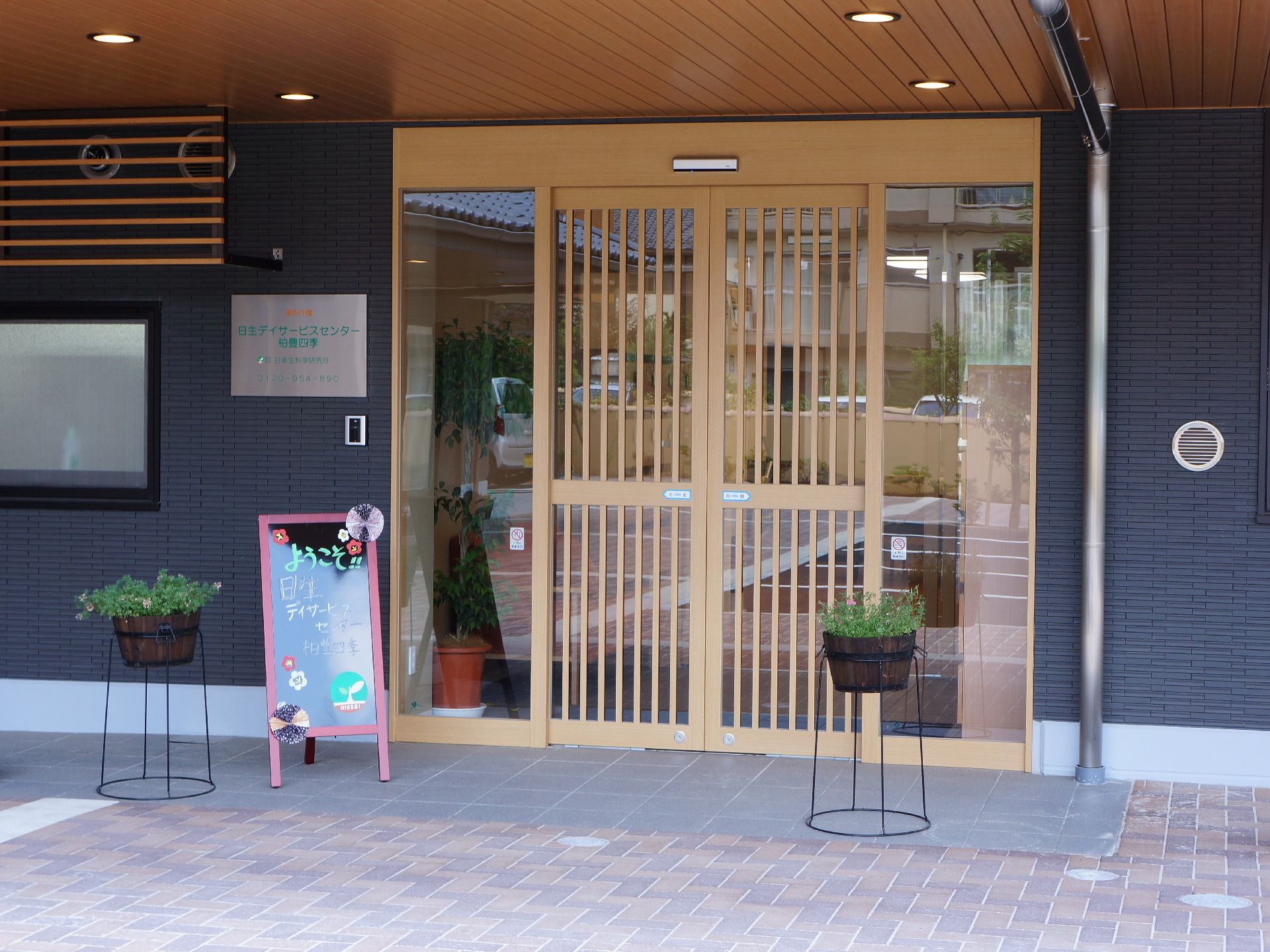 施設玄関の外壁は黒を基調としている。扉は木、屋根は瓦で作られている。玄関前には施設の看板と手書きで書かれた黒板が置かれている。