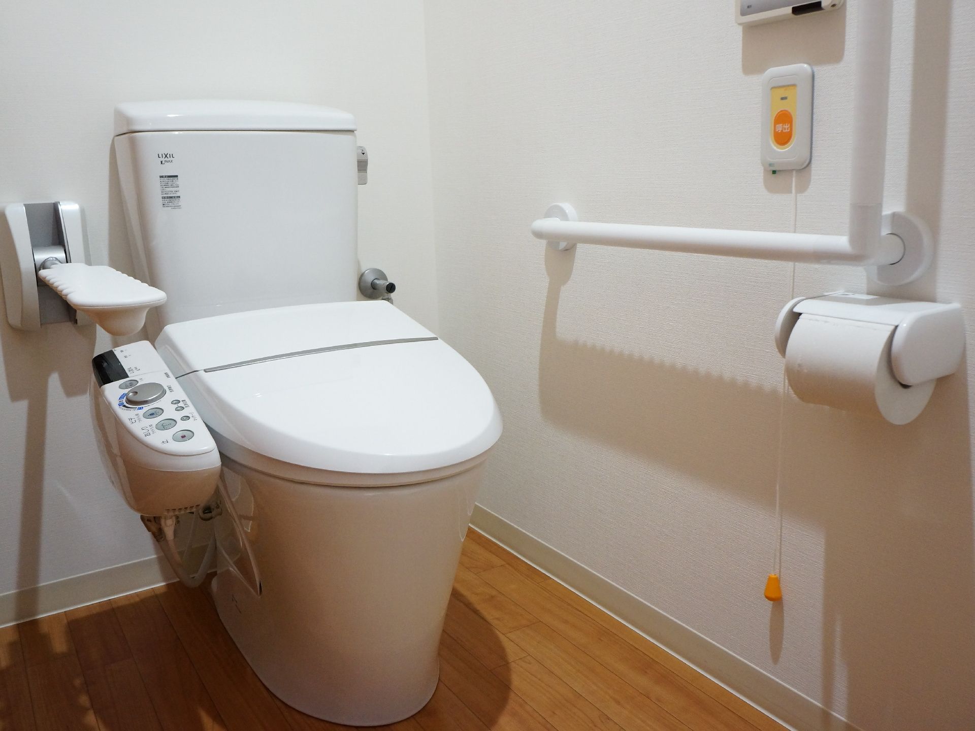 白色の壁に木目調の床のトイレスペース。便器横の壁にはエル型に手すりが設置されている。またナースコールが取り付けてある。