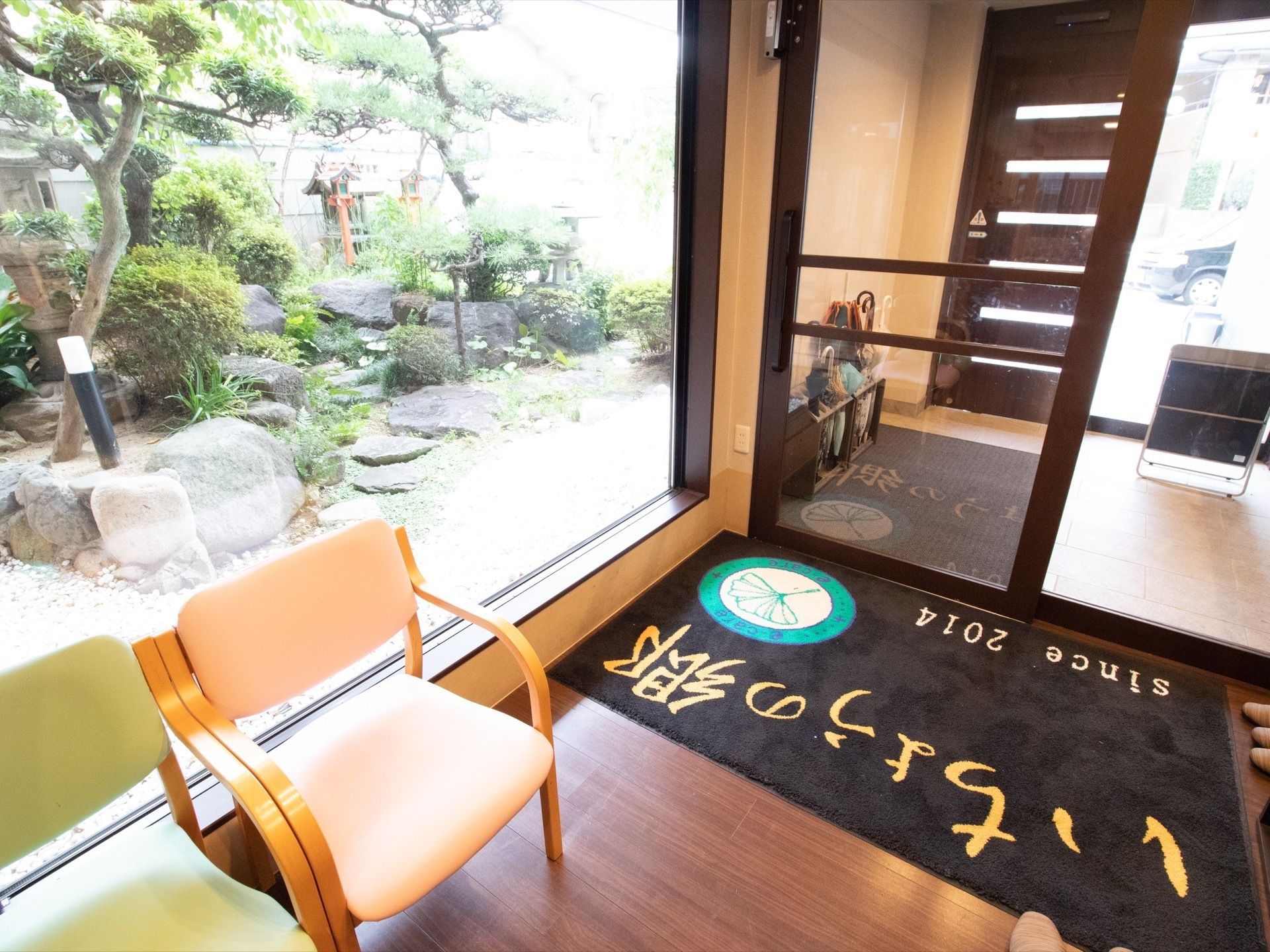 玄関扉は自動扉となっている。床に施設名の書かれた玄関マットが敷かれている。入口前に椅子が２脚置かれている。