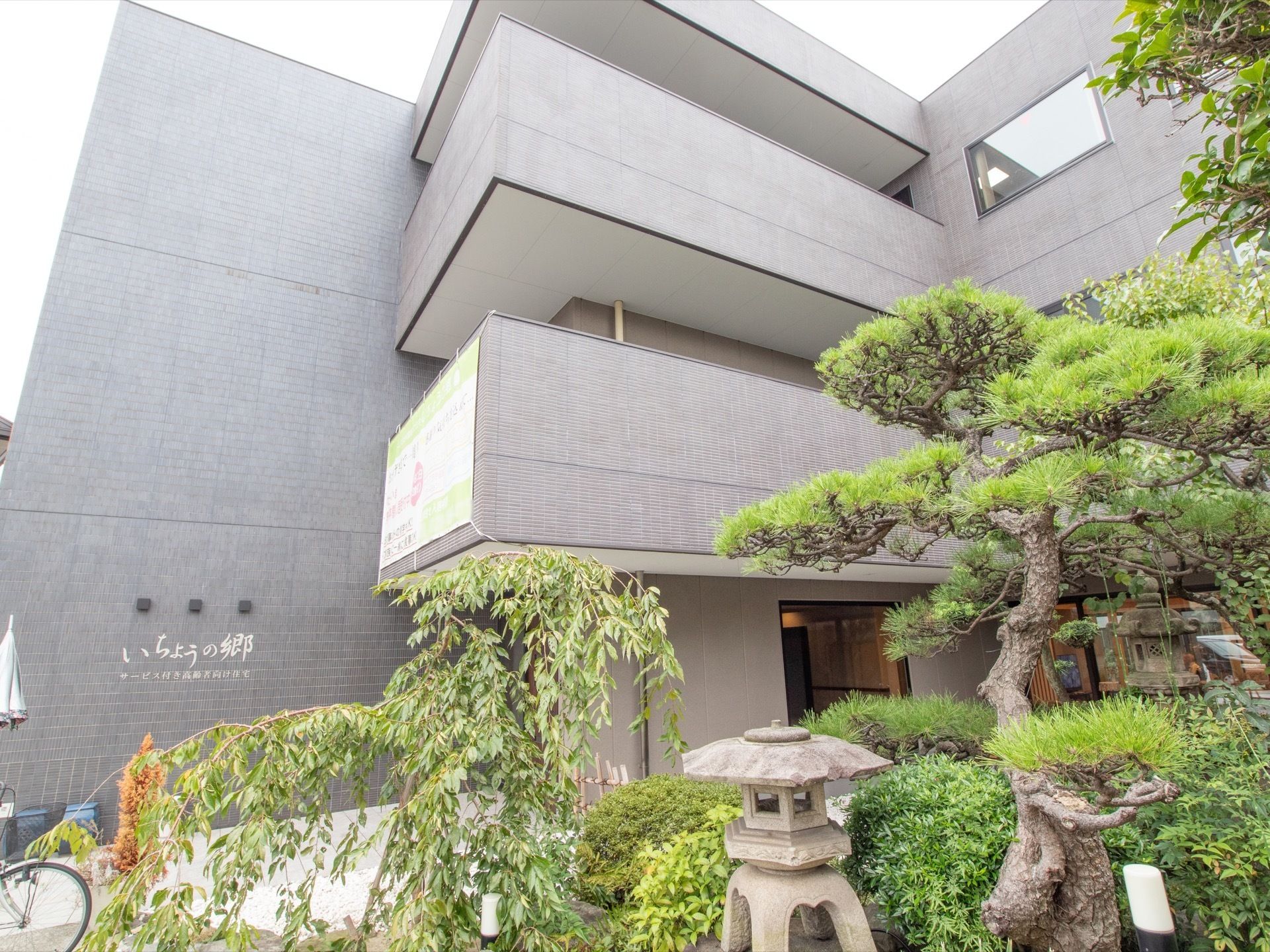 グレーの外壁の３階建ての建物である。壁に施設名が表示されている。手前に中庭が広がっている。自転車が停められている。