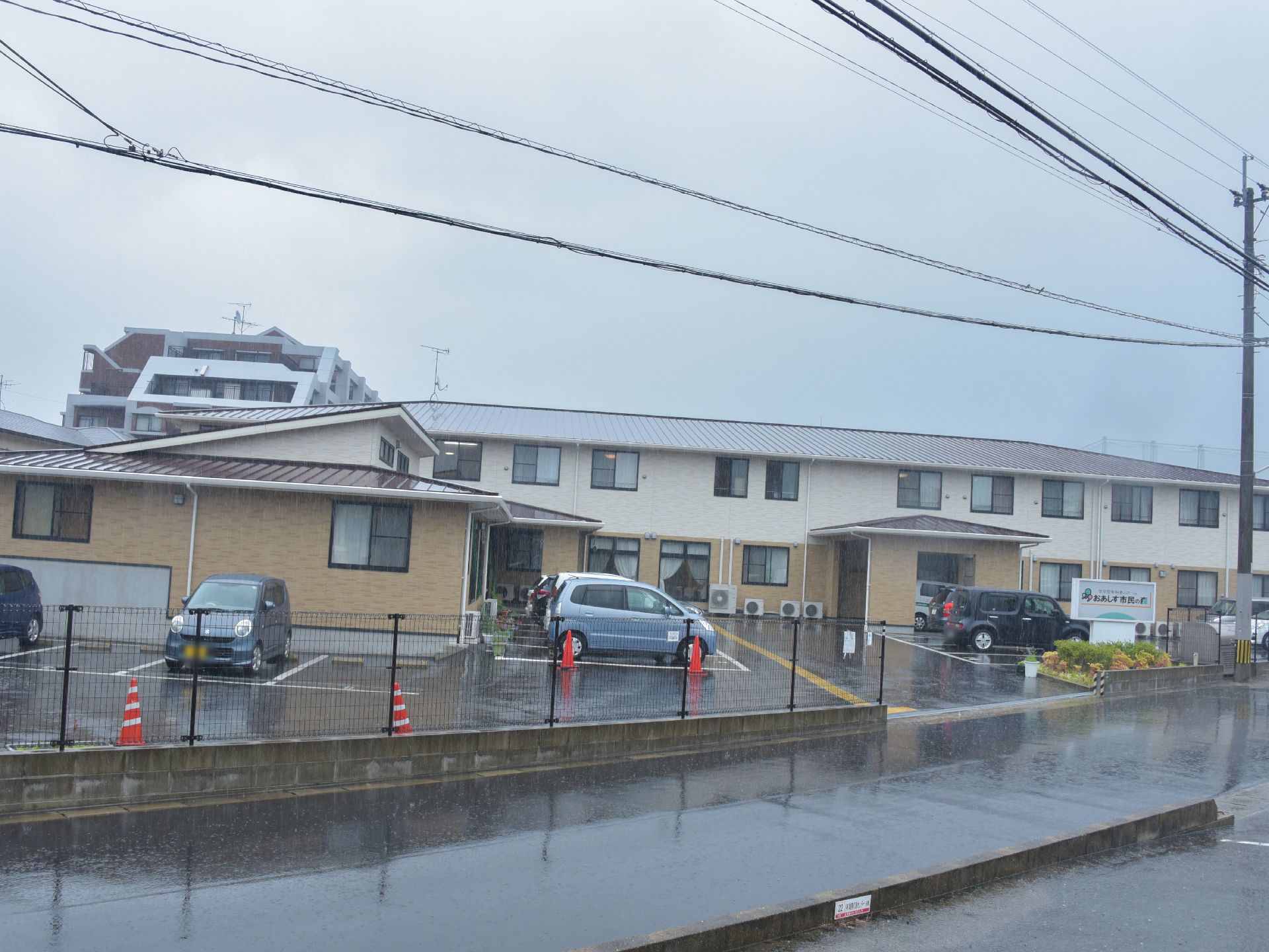 白と茶色の外壁の2階建てで目の前が駐車場の施設になっている。道路に面しており玄関には雨よけが張り出している。