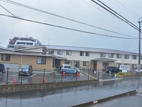 白と茶色の外壁の2階建てで目の前が駐車場の施設になっている。道路に面しており玄関には雨よけが張り出している。