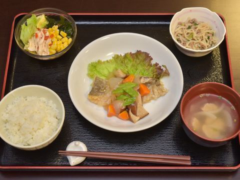 黒いトレイに健康を考えられた食事がのっている。野菜中心の主催が白い皿にのっており副菜が2品にご飯や味噌汁もついている。