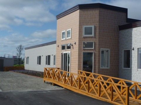 グレーと茶色のタイル貼りで、新築の建物の前に舗装された敷地がある。茶色い部分には玄関があり、５角形に飛び出している。木製のスロープがある。