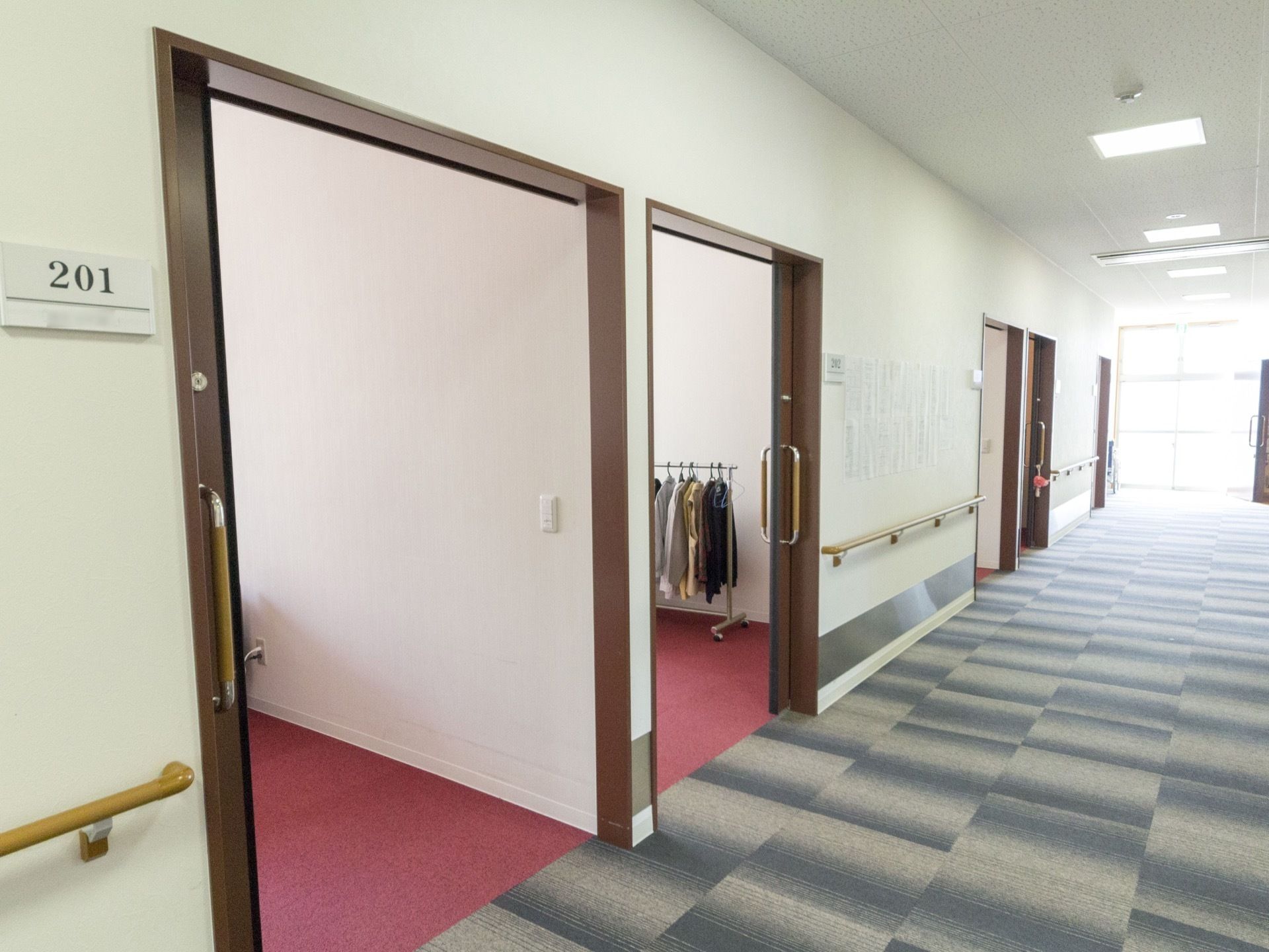 通路の床はブルー系の絨毯敷きである。居室の扉は引き戸であり、居室内の床はピンク色である。通路の壁には手すりが続いている。