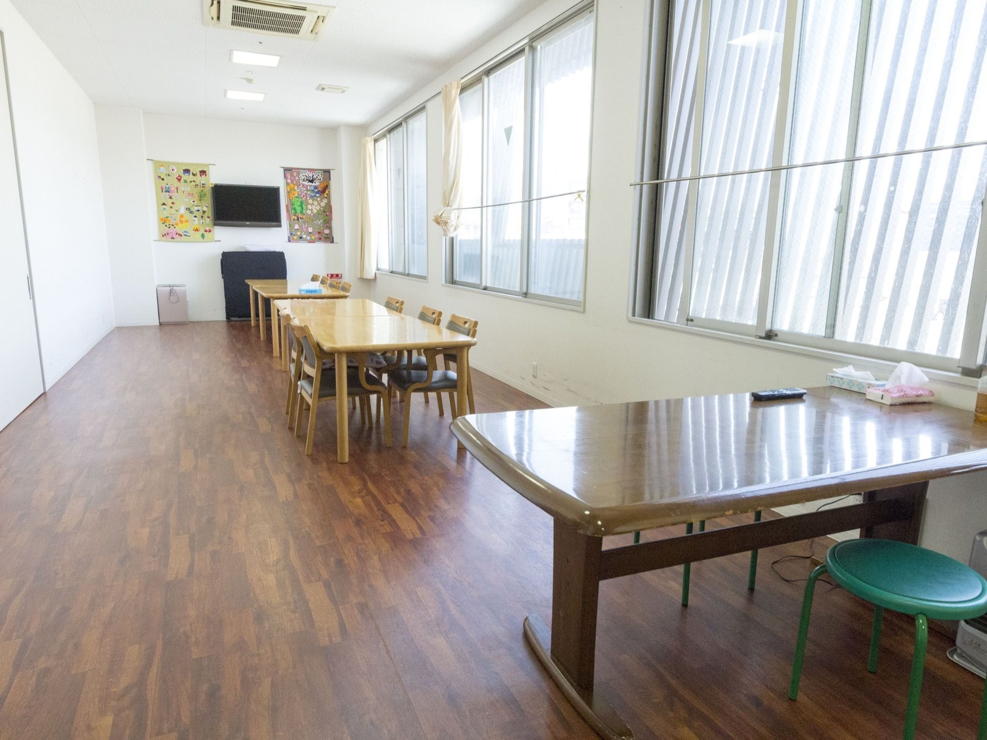 窓に面した横長の部屋である。壁にテレビがある。テーブルセットが窓に沿って2つある。天井にエアコンがある。
