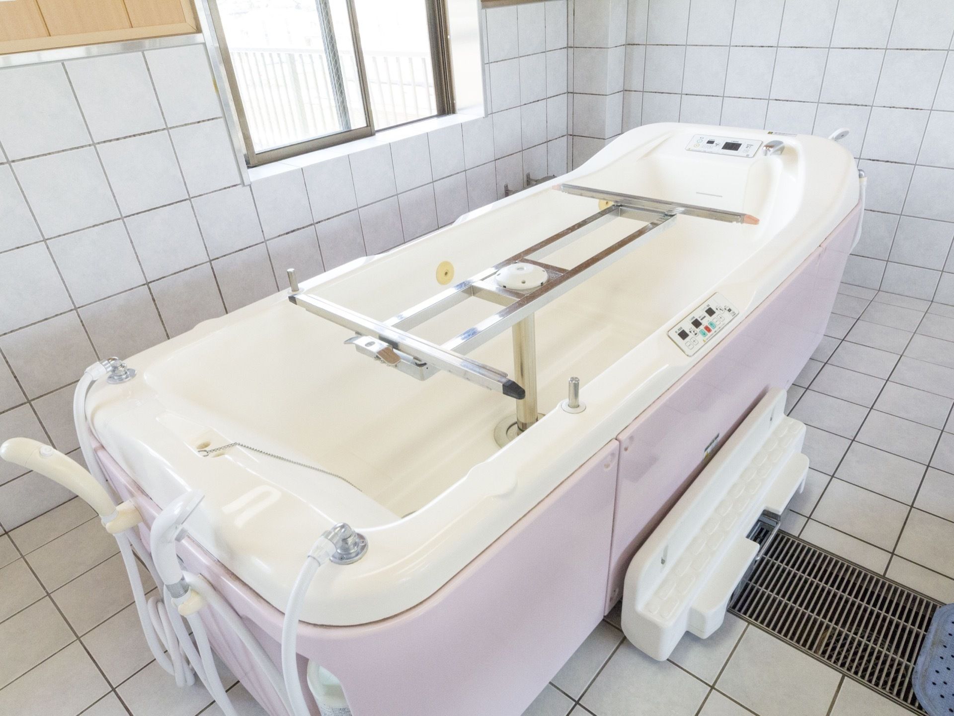 床と腰高まで白いタイルが貼られている。中央部に排水溝が続いており、上に介護浴槽が置かれている。寝たままでも利用できるタイプである。