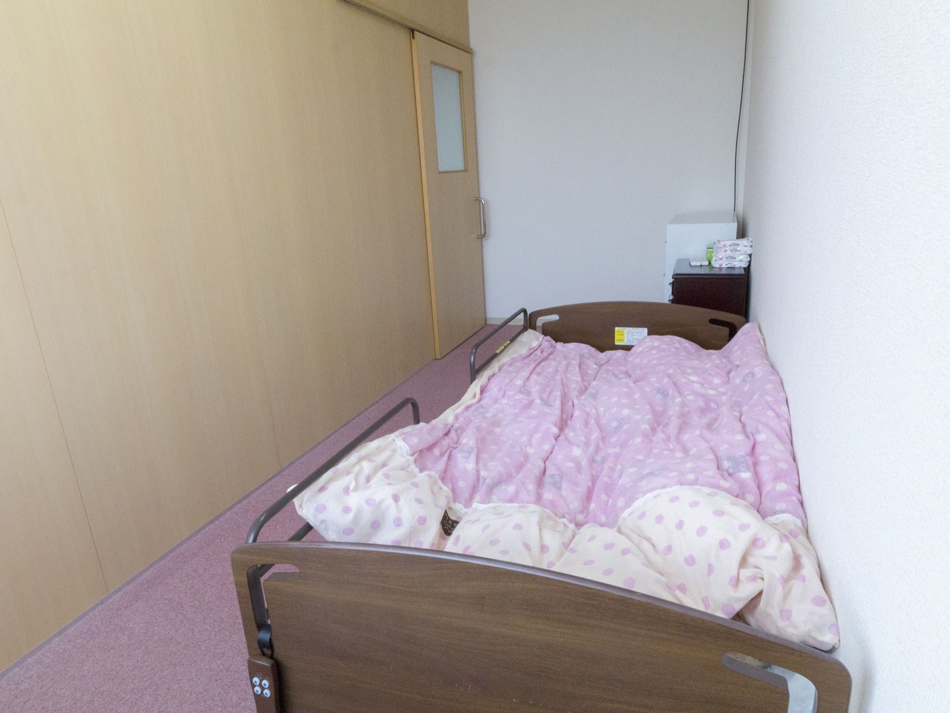 ドアはスライド式である。茶色の介護用の電動ベッドが設置されている。サイドレールがついている。床はピンク色のカーペットが敷かれている。