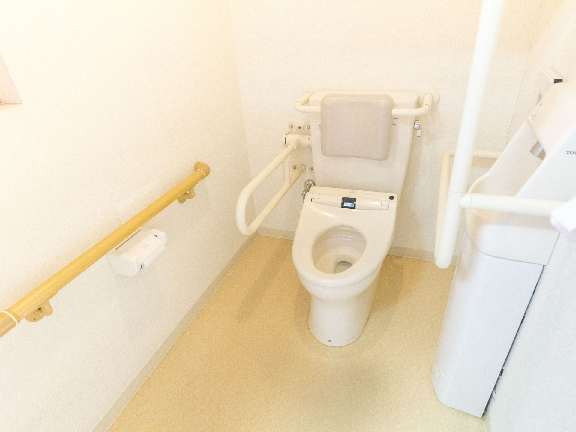 温水洗浄便座が設置されている。壁に手すりが完備されている。便座に背もたれと、可動式の手すりがついている。
