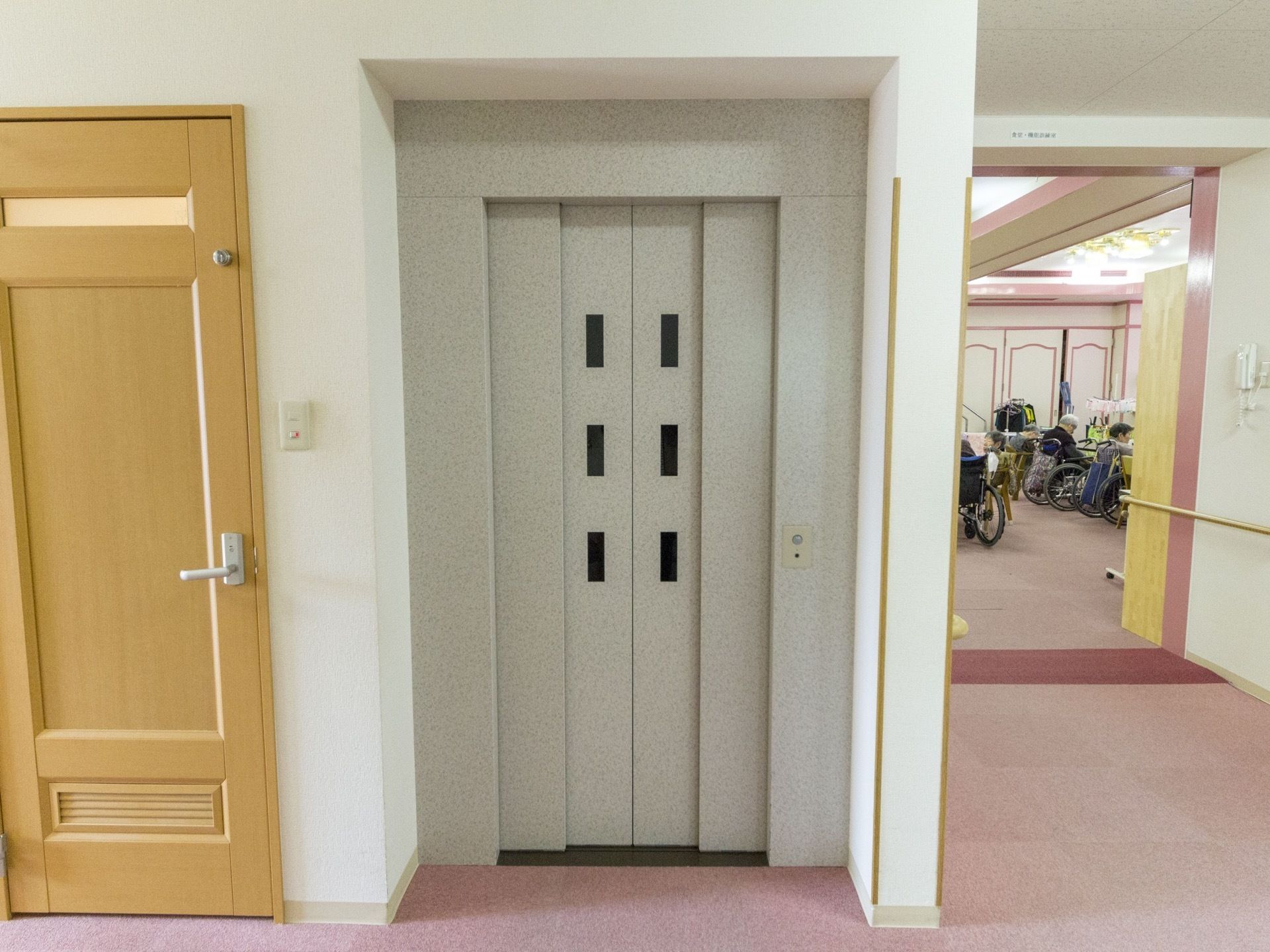 扉がグレーのエレベーターがある。周りの床はカーペット敷きになっており、足に負担が少なく移動がしやすい。
