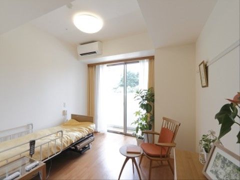 居室の床はフローリングになっており、介護用ベッド、テーブル、手すり付きの椅子、観葉植物などがおかれている。
