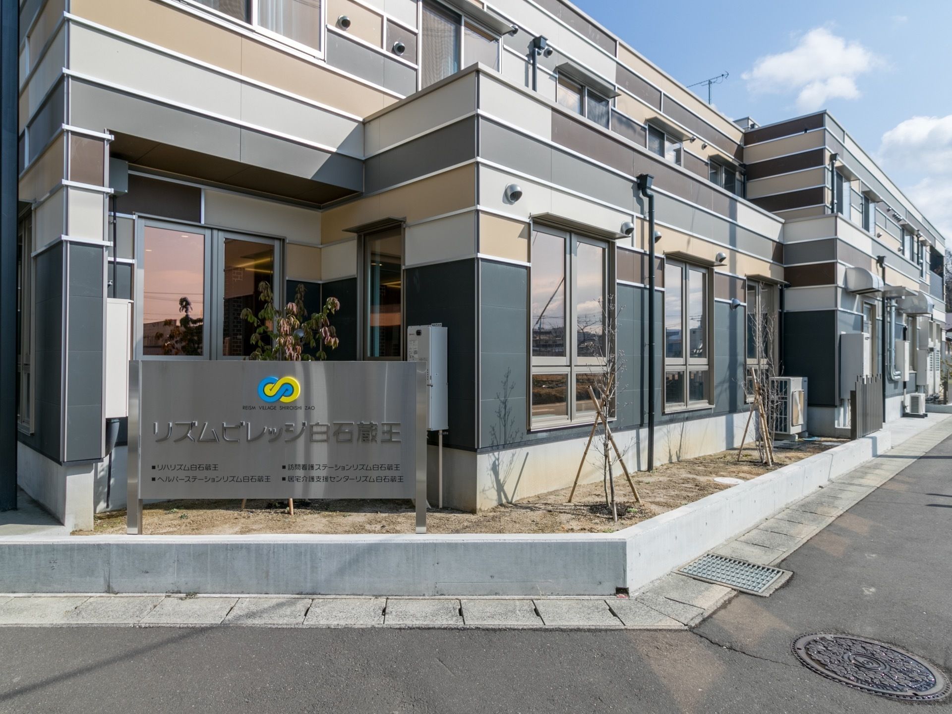 2階建てで道路に面した立地になっている。窓がたくさんあり、施設内が明るい空間になっている。施設の周りには木々が植えられている。