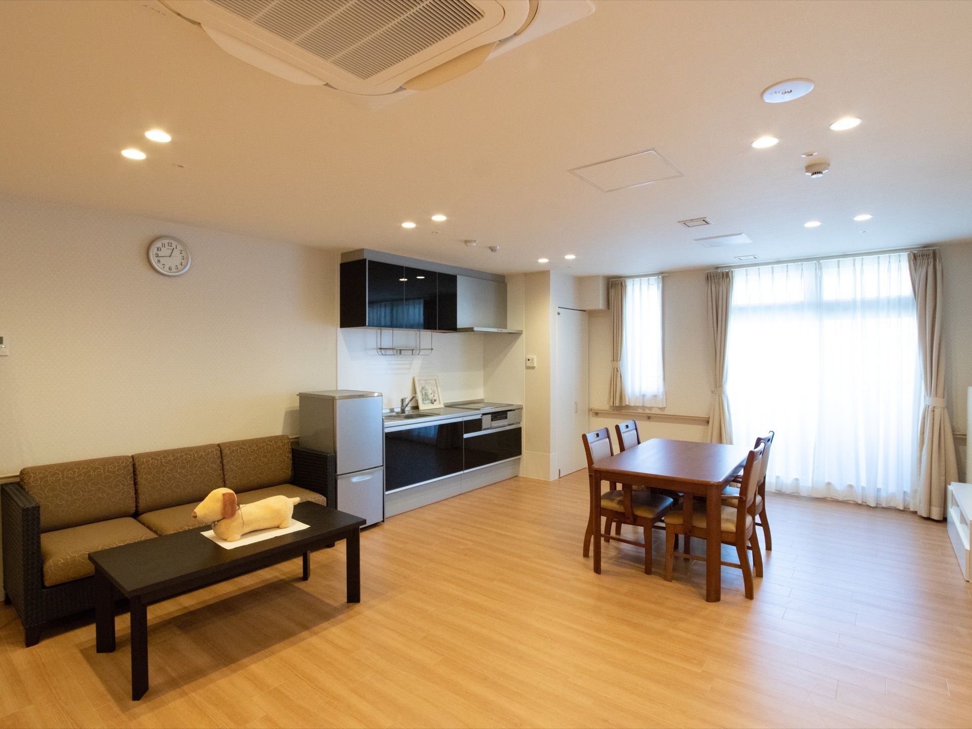 日当たり良好な広い部屋にシステムキッチンと冷蔵庫、ダイニングセット、テーブルとソファが用意されている。
