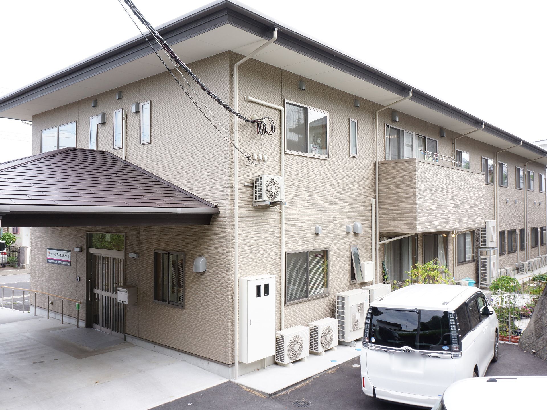 施設は2階建ての建物で、上階には部分的にベランダが設置されている。左側の玄関の上には大きな茶色い屋根が付いている。
