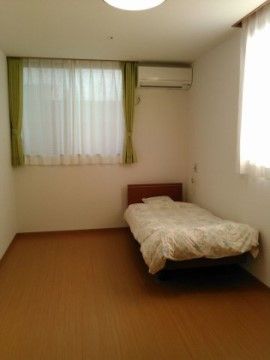 居室にはエアコンが設置され窓があり、壁と天井の色は白色系で、明るい雰囲気である。床はフローリングになっており、ベッドが置かれている。