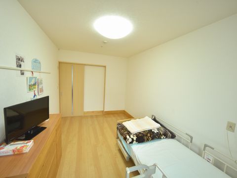 白色の壁と天井に木目の床の内装の居室である。引き戸のドア、壁際には介護用ベッド、反対壁際には収納家具がある。その上にはテレビが置かれている。
