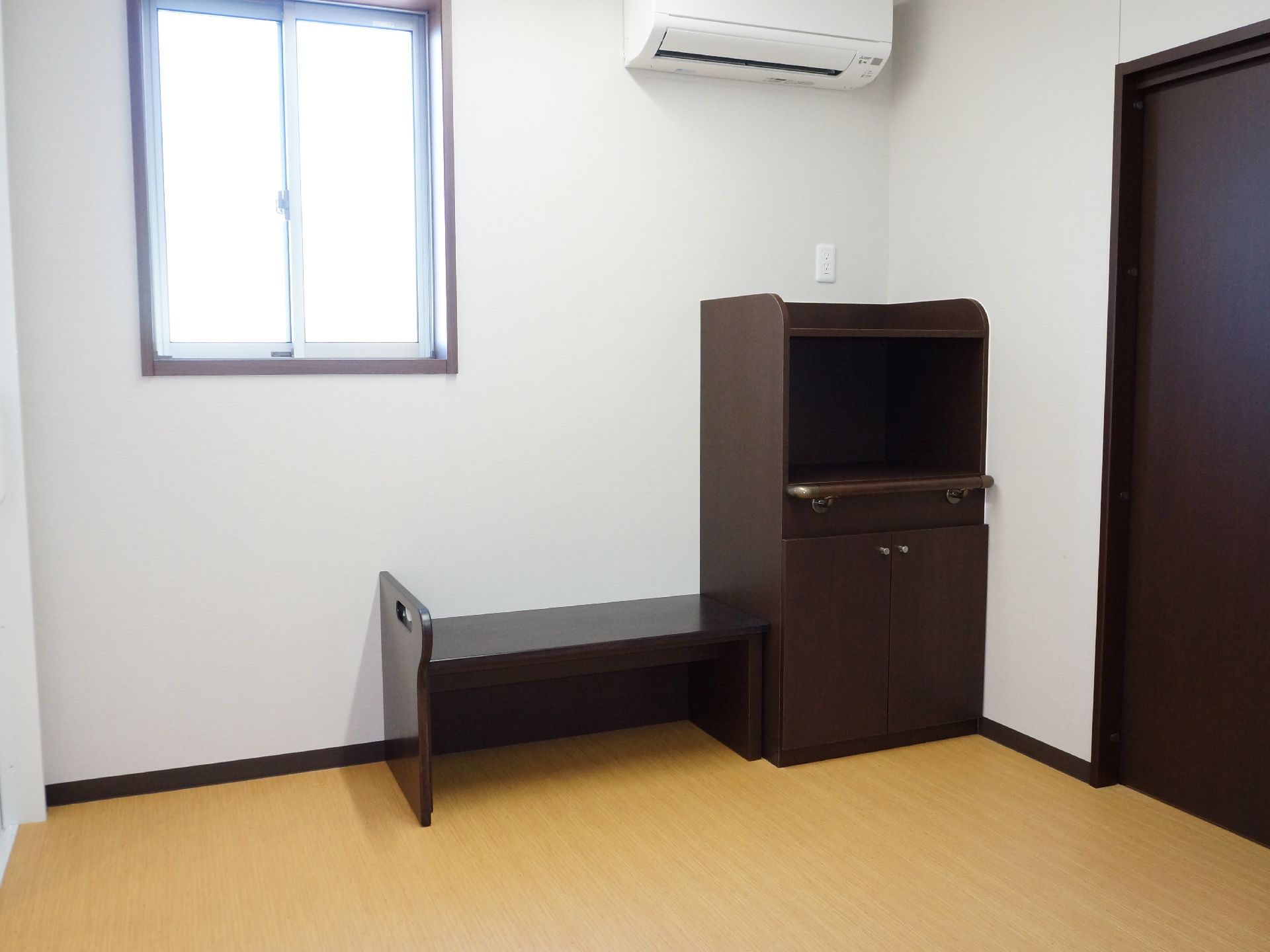 焦げ茶のドアに、白色の壁と黄土色の床である。天井のすぐ下にエアコンが設置されている。縦長の窓があり、焦げ茶色の腰かけ台と収納家具がある。