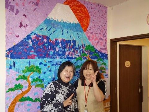富士山を象った装飾の壁を背に仲睦まじい入居者とヘルパー