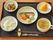 ご飯と汁物、焼いた鮭とサトイモや人参等の野菜、赤や黄色のパプリカが入った煮物、オレンジなどが、黒いトレーに並べられている。