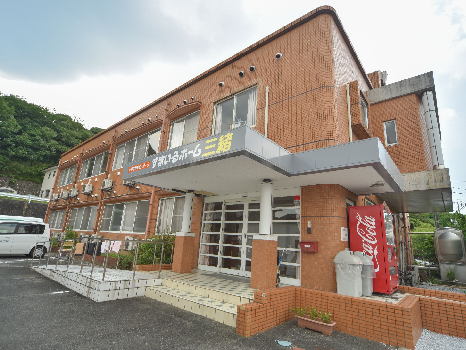 外観は、全体的に茶色系でまとめられたデザインになっている。玄関横の壁にはポストがあり、その向こうには自動販売機もある。