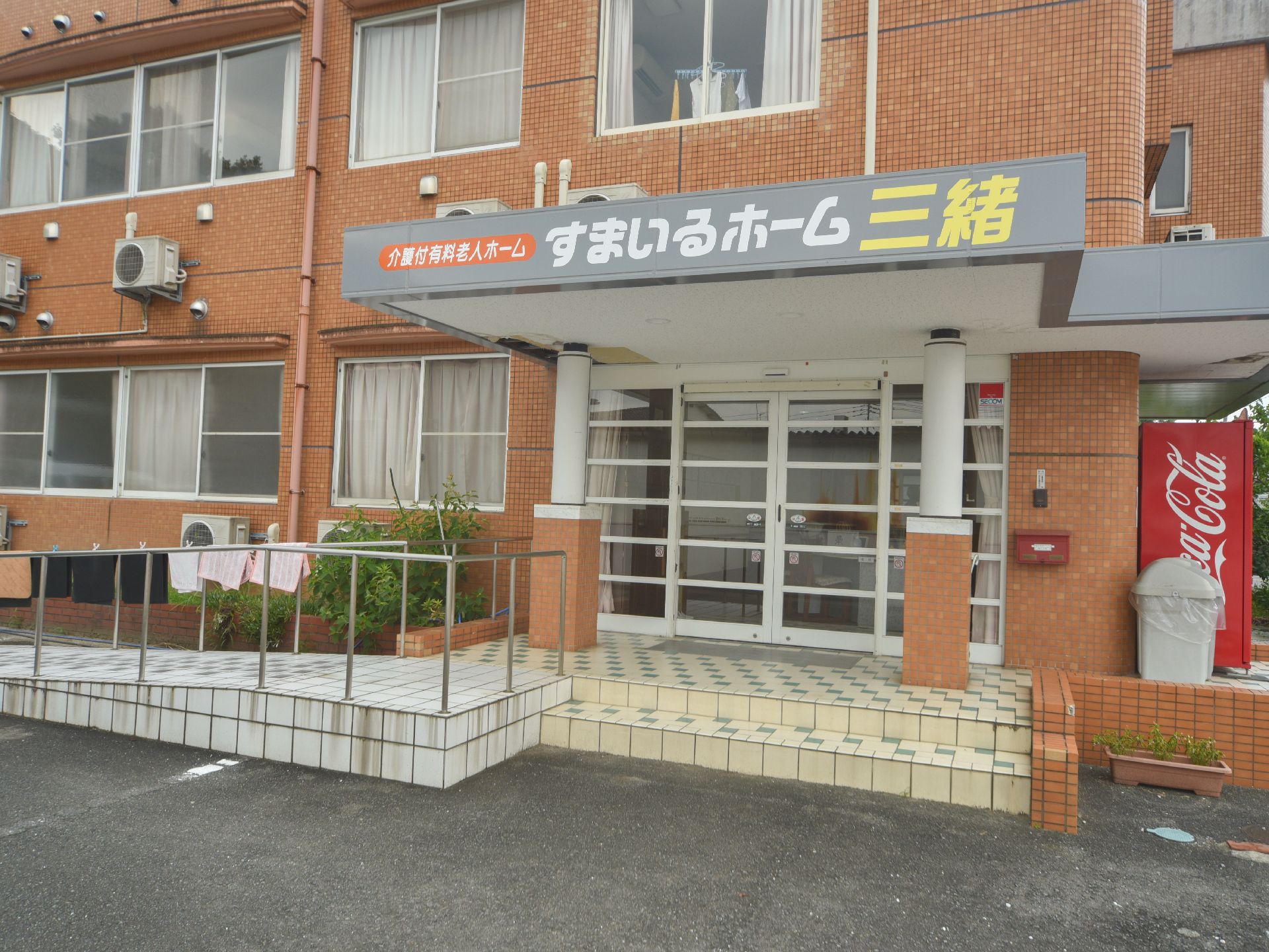 白い屋根が張り出しているスペースが、玄関前に設置されている。屋根の正面には、大きな文字で施設名が書かれている。