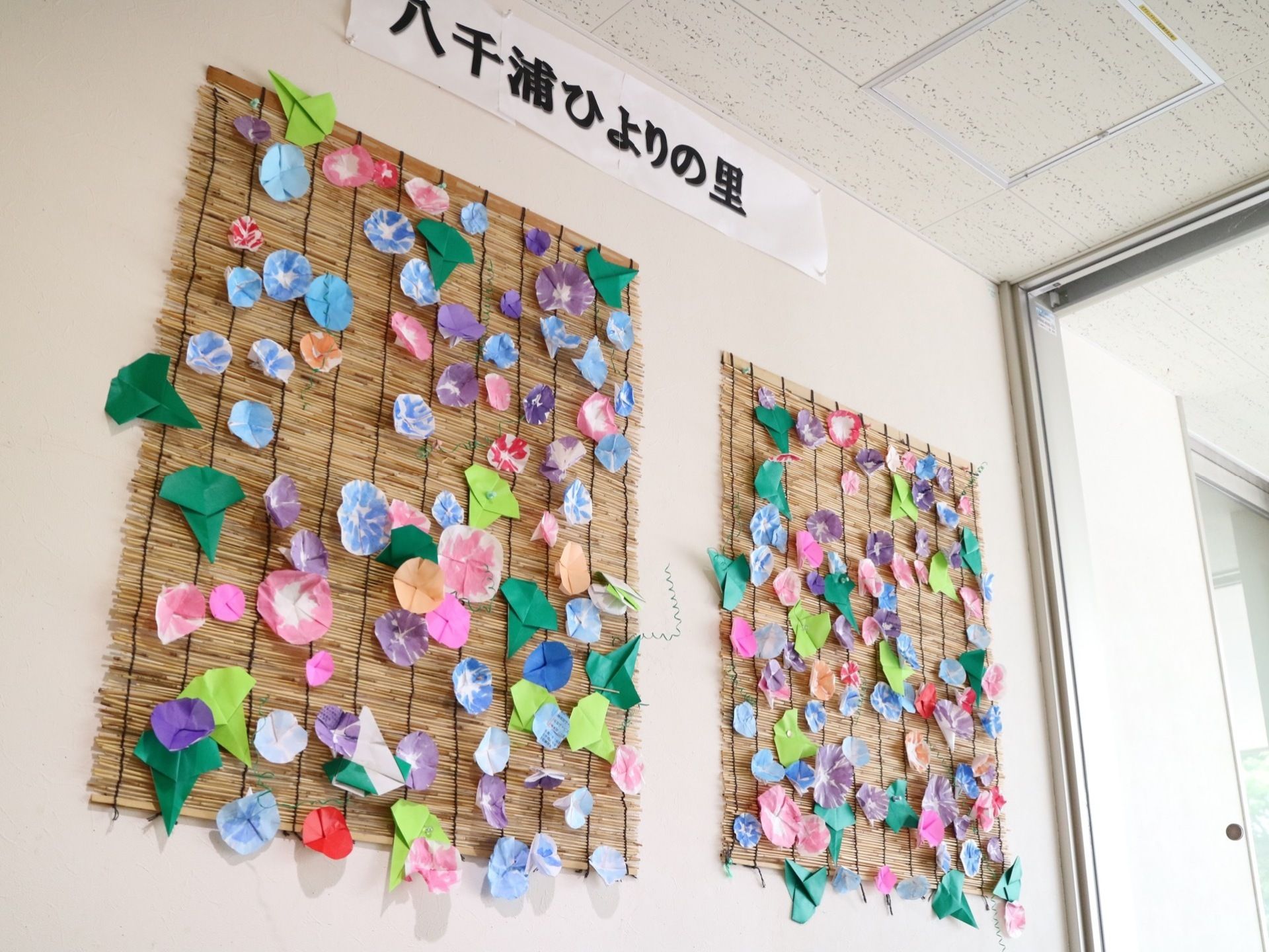 入居者様が紙で作られた季節を代表する花の作品が、すだれに貼られてたくさん飾られている。壁の上の方に施設名称が書かれている。