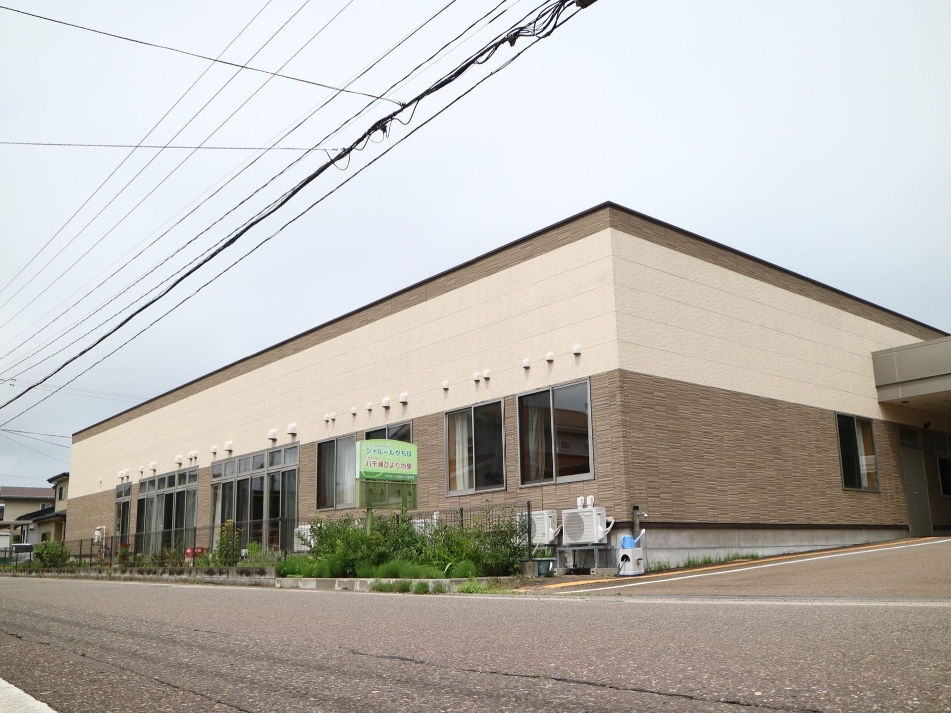 茶系統の色とベージュ色のツートンカラーの外壁を持つ、平屋建ての施設である。道路沿いに緑色の看板が設置されている。