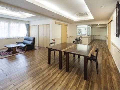 空間にゆとりがあり、車椅子が１台置かれている。通路には手すりが設置されており、部屋の真ん中にはテーブルと椅子がセットで用意されている。