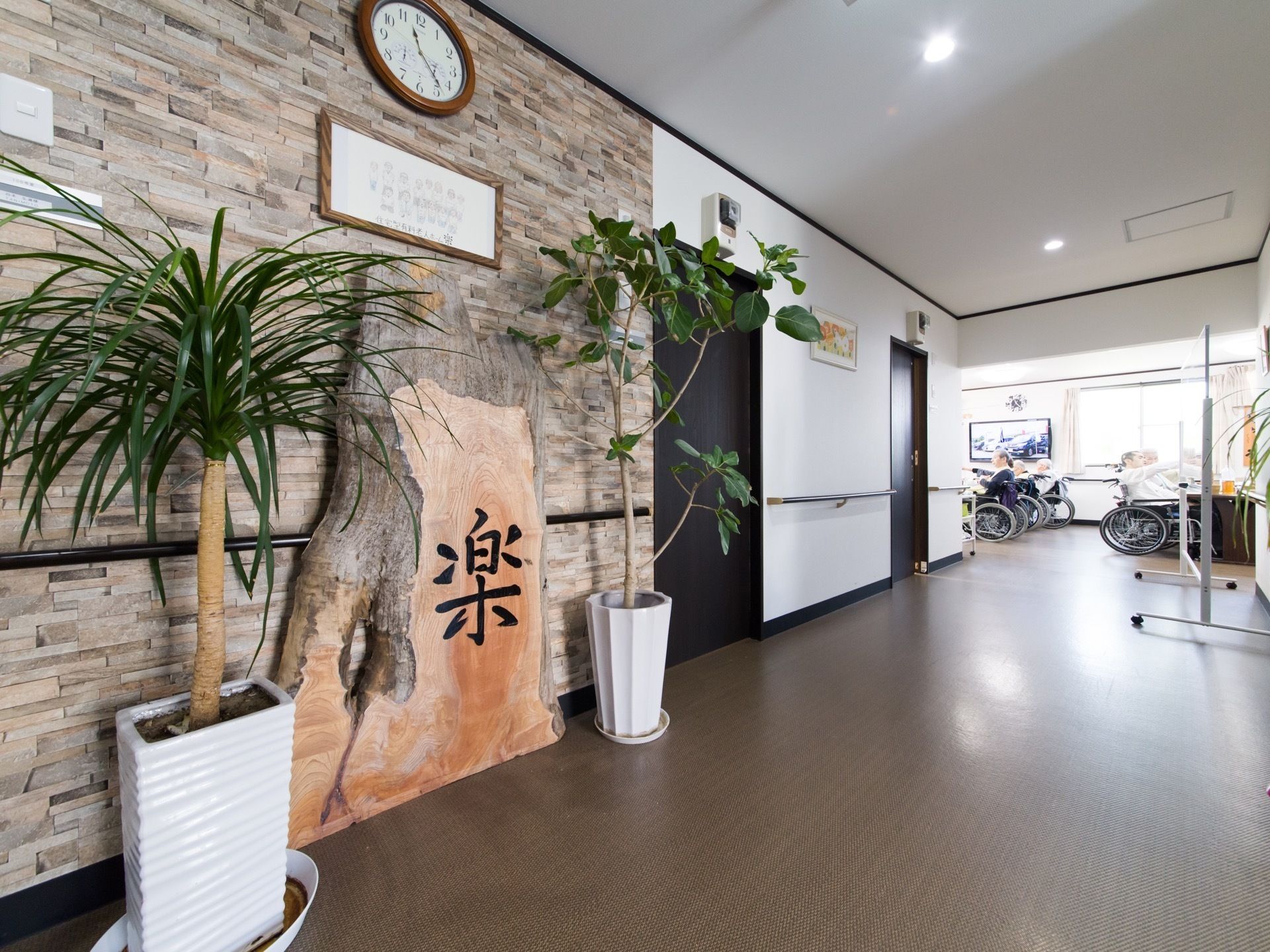 通路の壁には、時計、施設名の入った木の看板、観葉植物が設けられている。奥には車椅子の入居者様が集まっている。