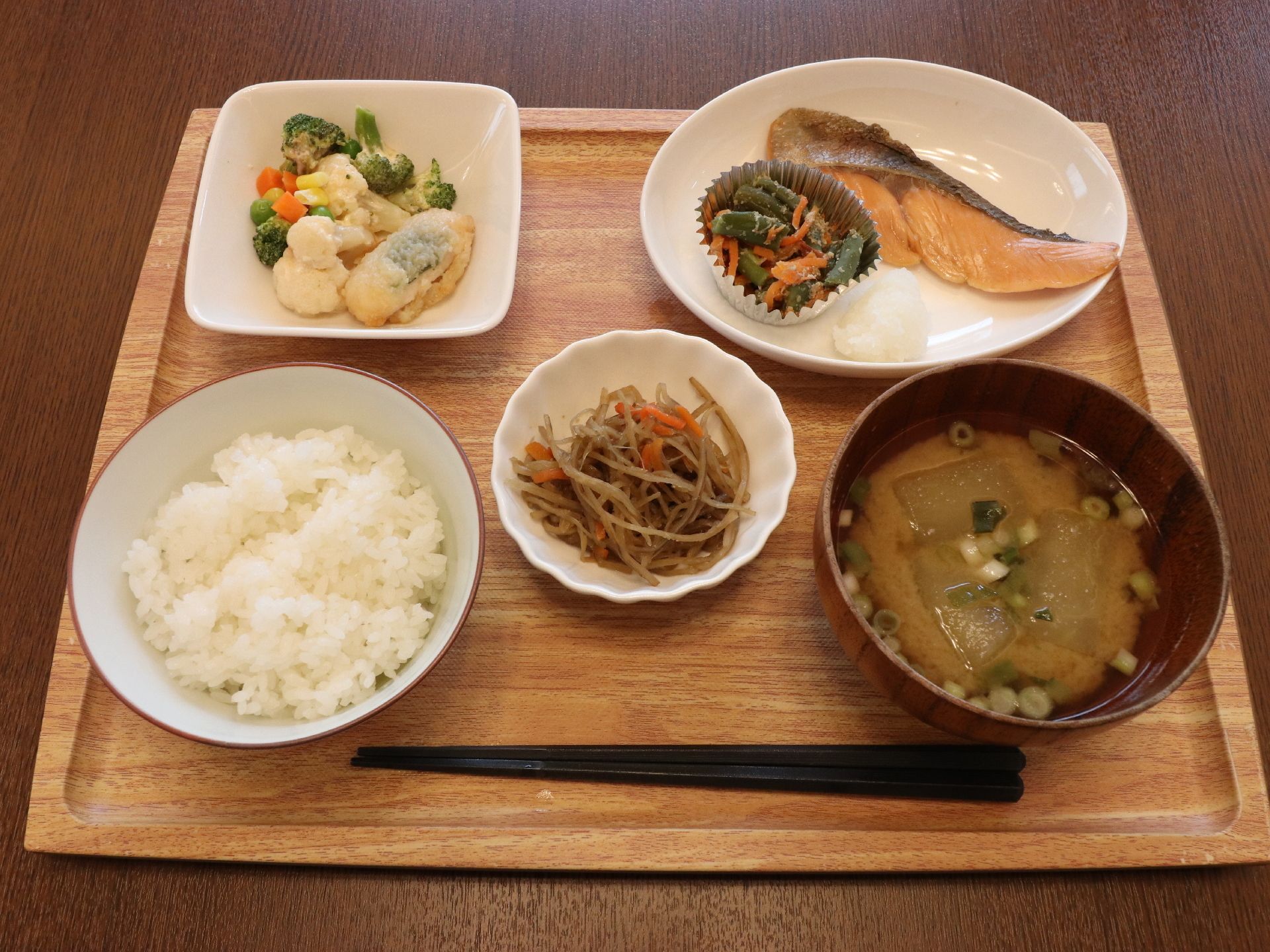 ご飯と汁物、ゴボウとニンジンのきんぴら、焼いた鮭と大根おろし、ブロッコリーなどのサラダが、茶色のトレーに並んでいる。