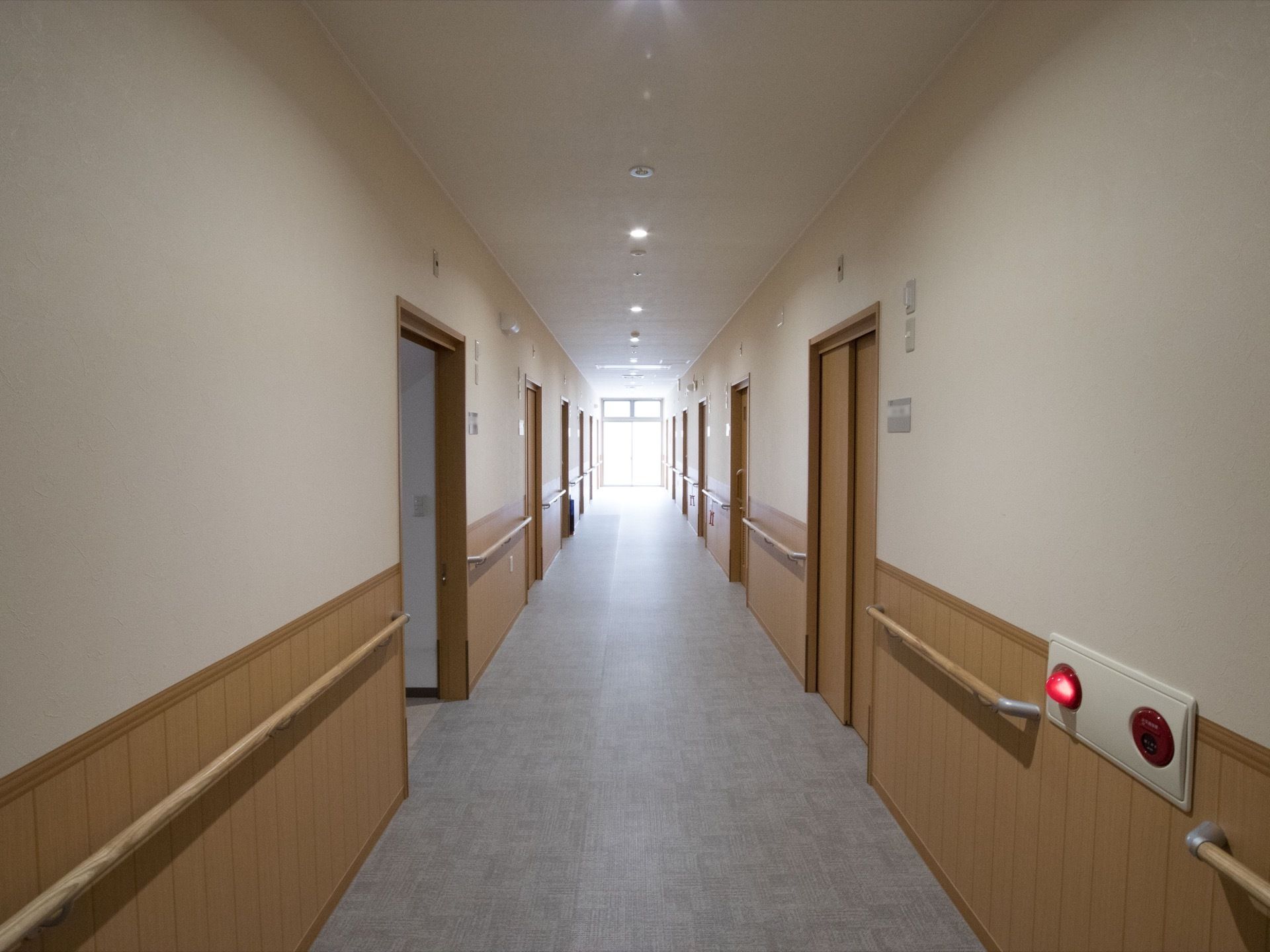 廊下の両側に居室への扉が等間隔で並んでいる。ドアの横にネームプレートがあり、壁に手すりが設置されている。