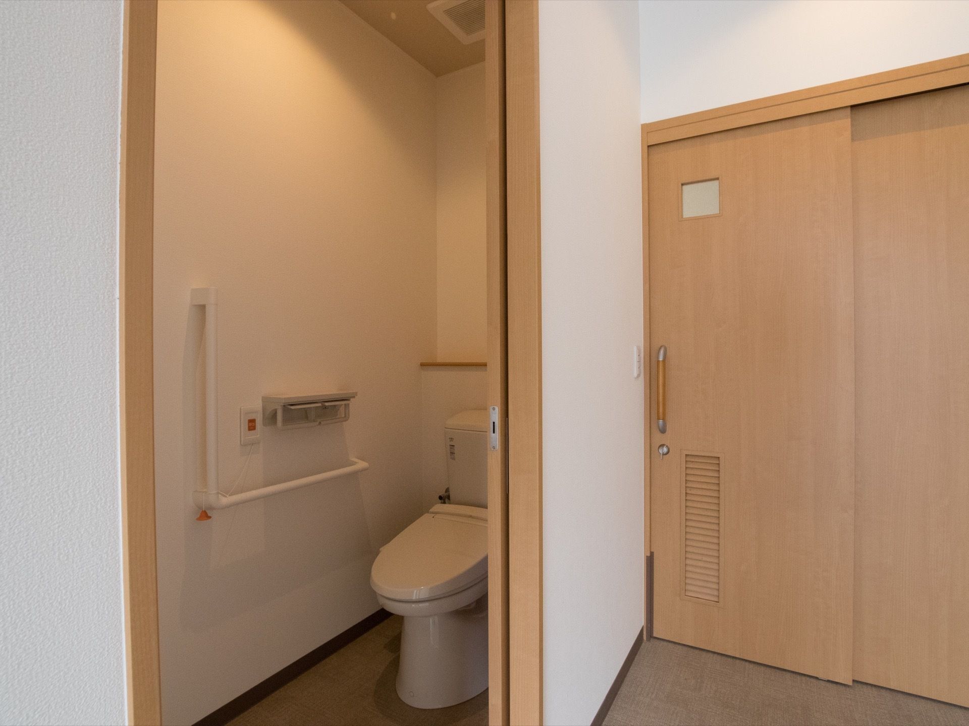 居室の入口の横にトイレがある。壁にはＬ字型の手すりが取りつけされており、トイレットペーパーが２か所にある。