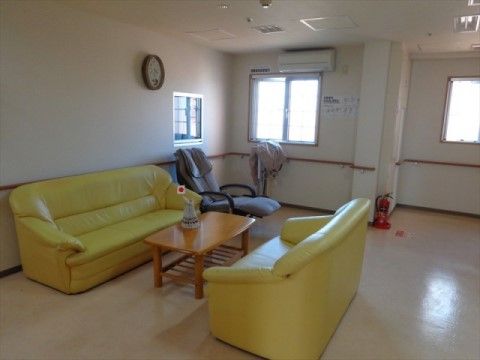 白い壁に、茶色の手すりが設置された広々とした空間。黄色が鮮やかなソファセットとマッサージチェアが設置されている。