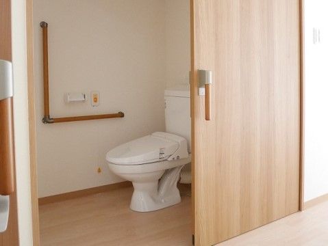 温水洗浄便座付きのトイレ。白い壁には手すりが付いており、ナースコールが設置されている。床と扉は明るい木目で、扉はスライド式になっている。