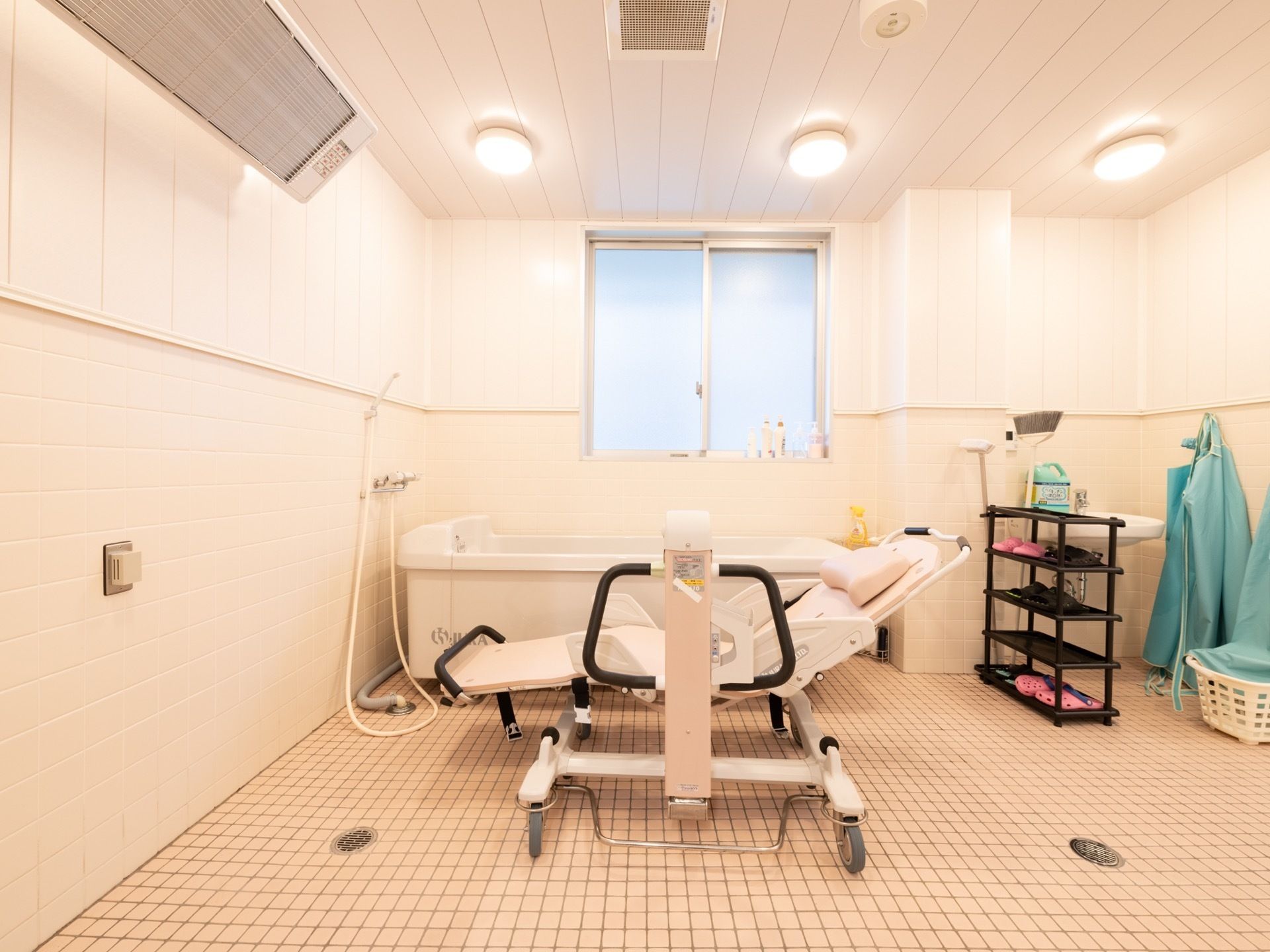 介助用の入浴器具が置かれている。広で統一されており、広く開放的である。清掃用品が棚に置かれている。防水エプロンが壁に掛けられている。