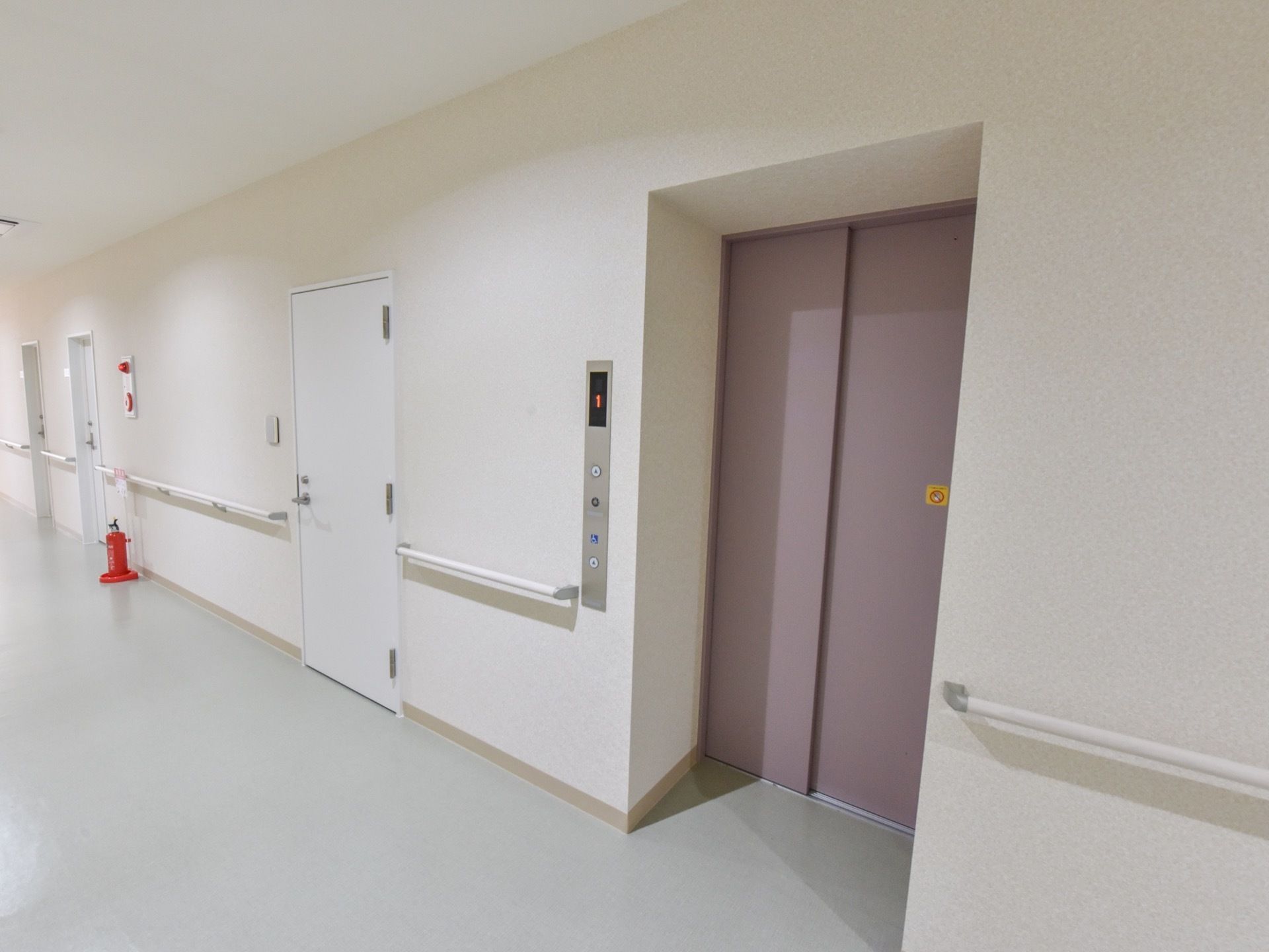 薄紫色のドアが取り付けられたエレベーターである。左側の壁に非常ベルが設置されており、すぐ下に消火器が置いてある。